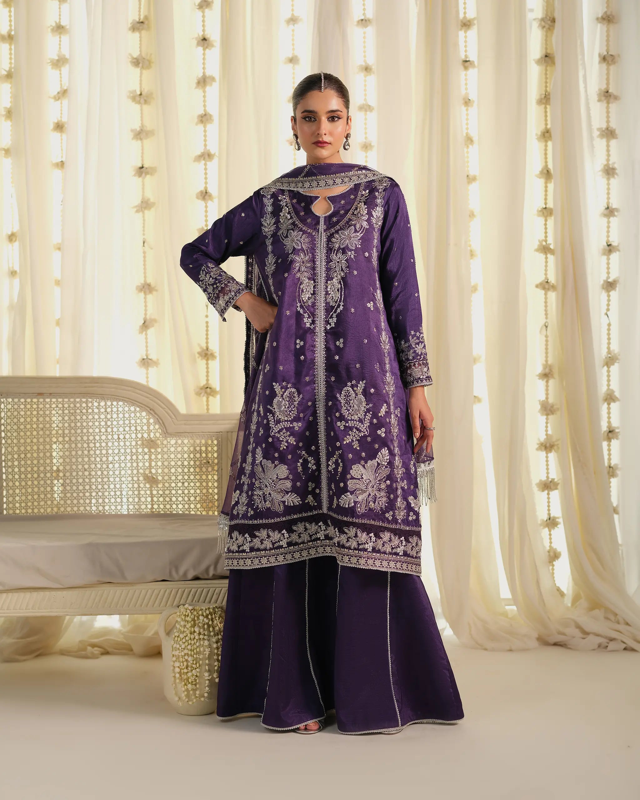 Model stands in front of a white chaise lounge wearing a deep purple 'kameez﻿' and flared trousers with silver embroidery from the 'Pehla Khat Khumaar﻿' collection.