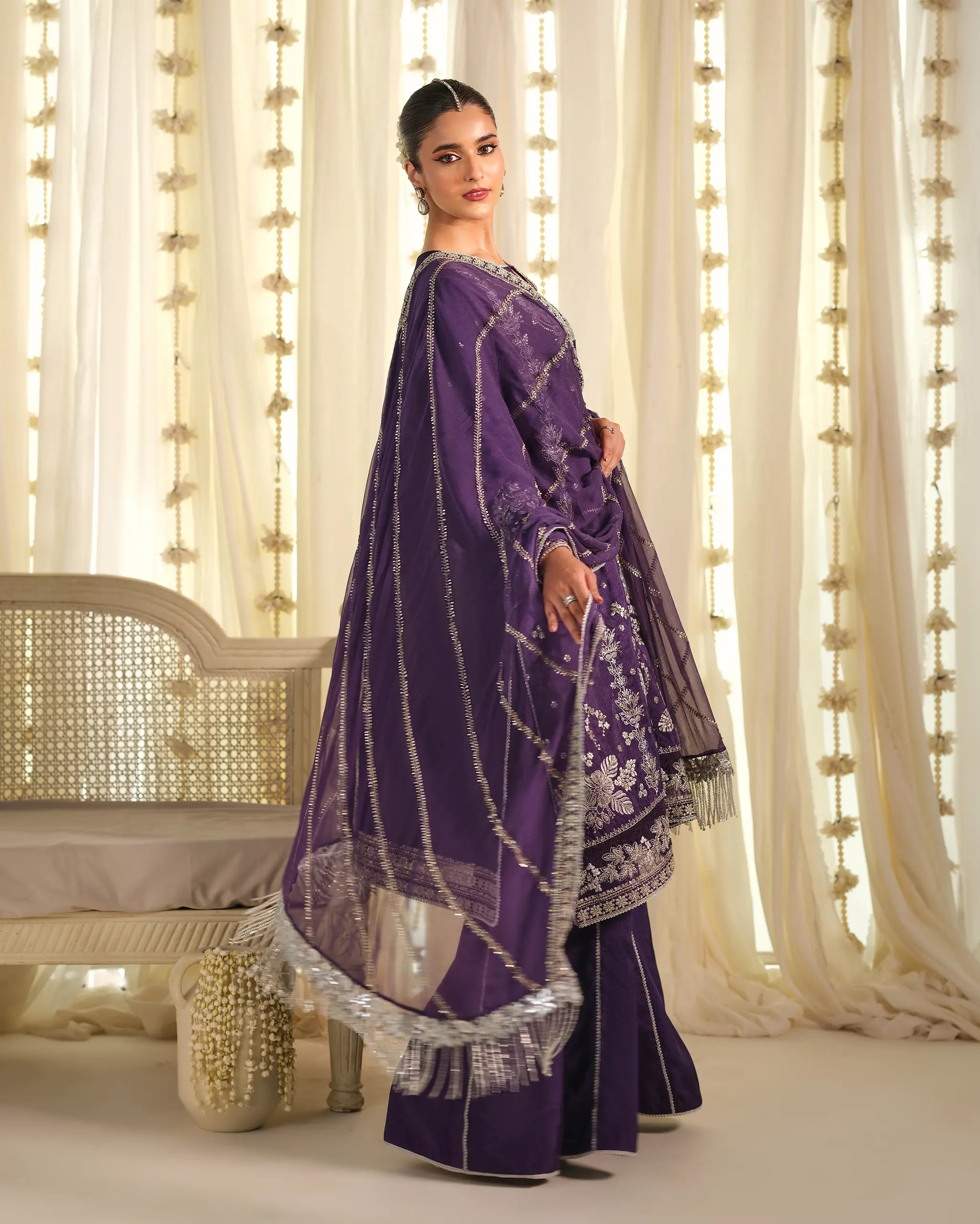 A three-quarter view of a model in the purple embroidered 'Khumaar﻿' suit, looking over her shoulder with the silver-fringed organza 'dupatta﻿' displayed.
