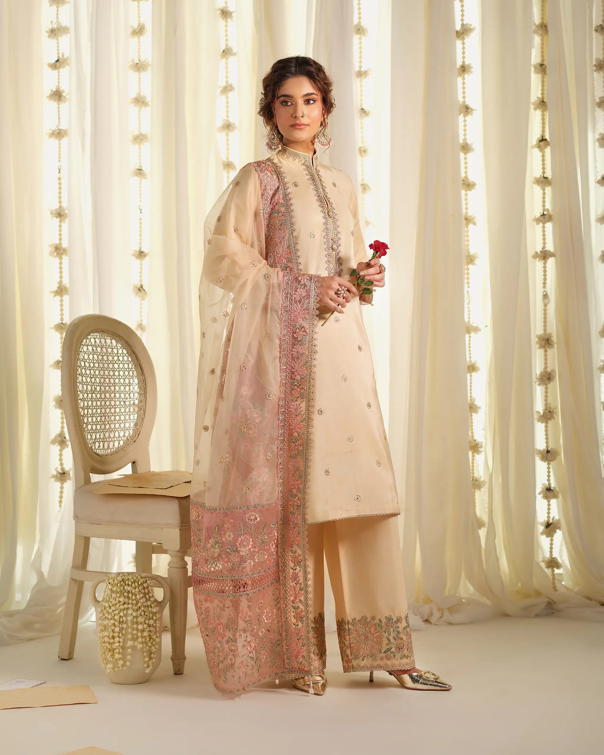 Model stands holding a red rose in a raw silk suit 'Tehreer', from the 'Pehla Khat﻿' collection, featuring a pink embroidered organza dupatta﻿.