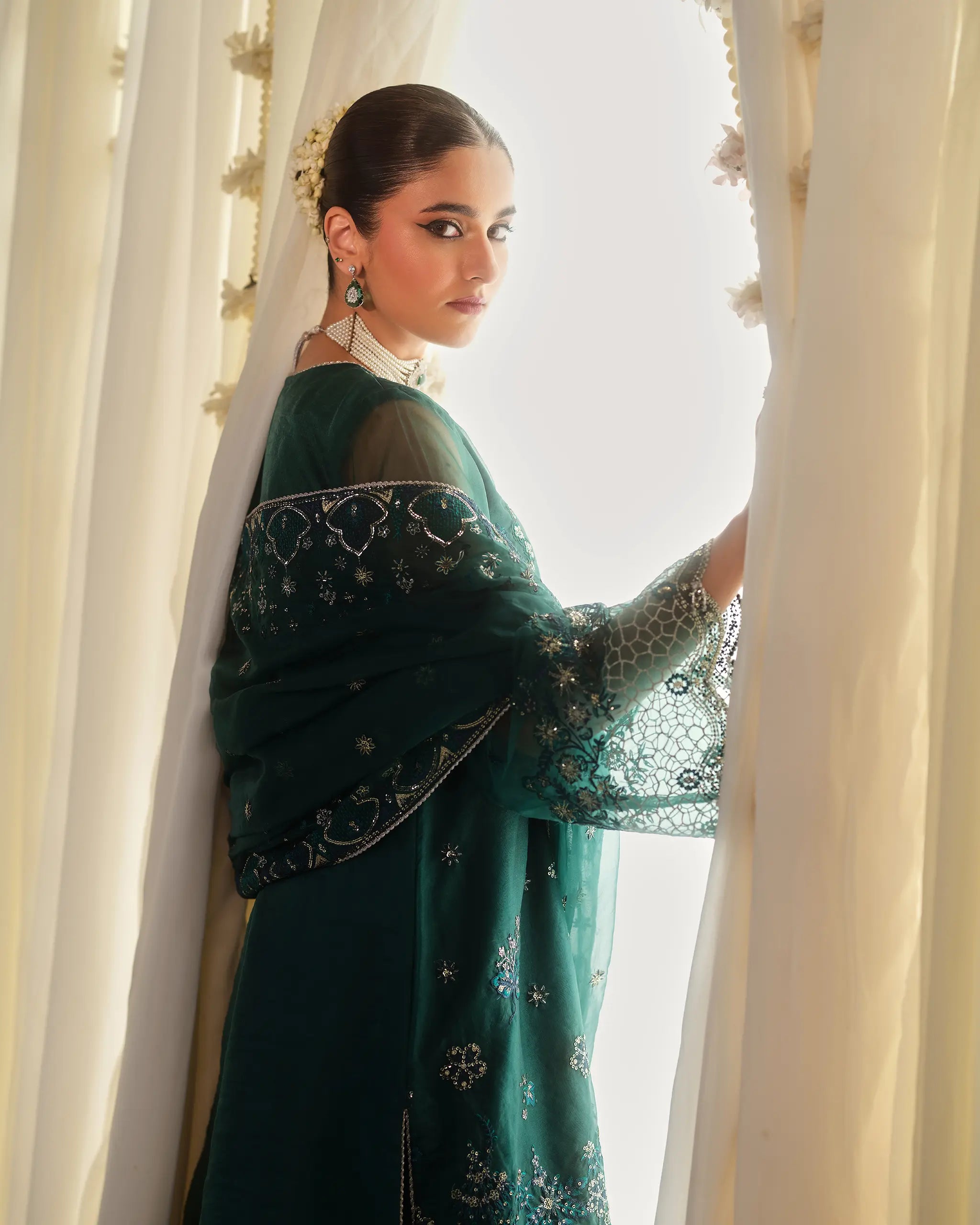 Model looking over her shoulder while parting a curtain, dressed in the emerald green 'Vasl﻿' suit with detailed embroidery on the sleeves and dupatta﻿