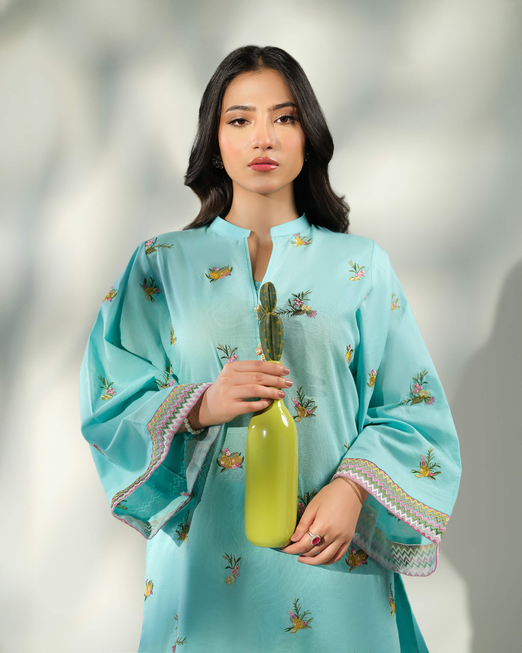 Sinoor Shop pret casual 2025 Amal Light Blue outfit. A woman holds a small green vase, wearing a light blue kurta with floral and bird embroidery on flared sleeves. The cuffs have a geometric pattern.