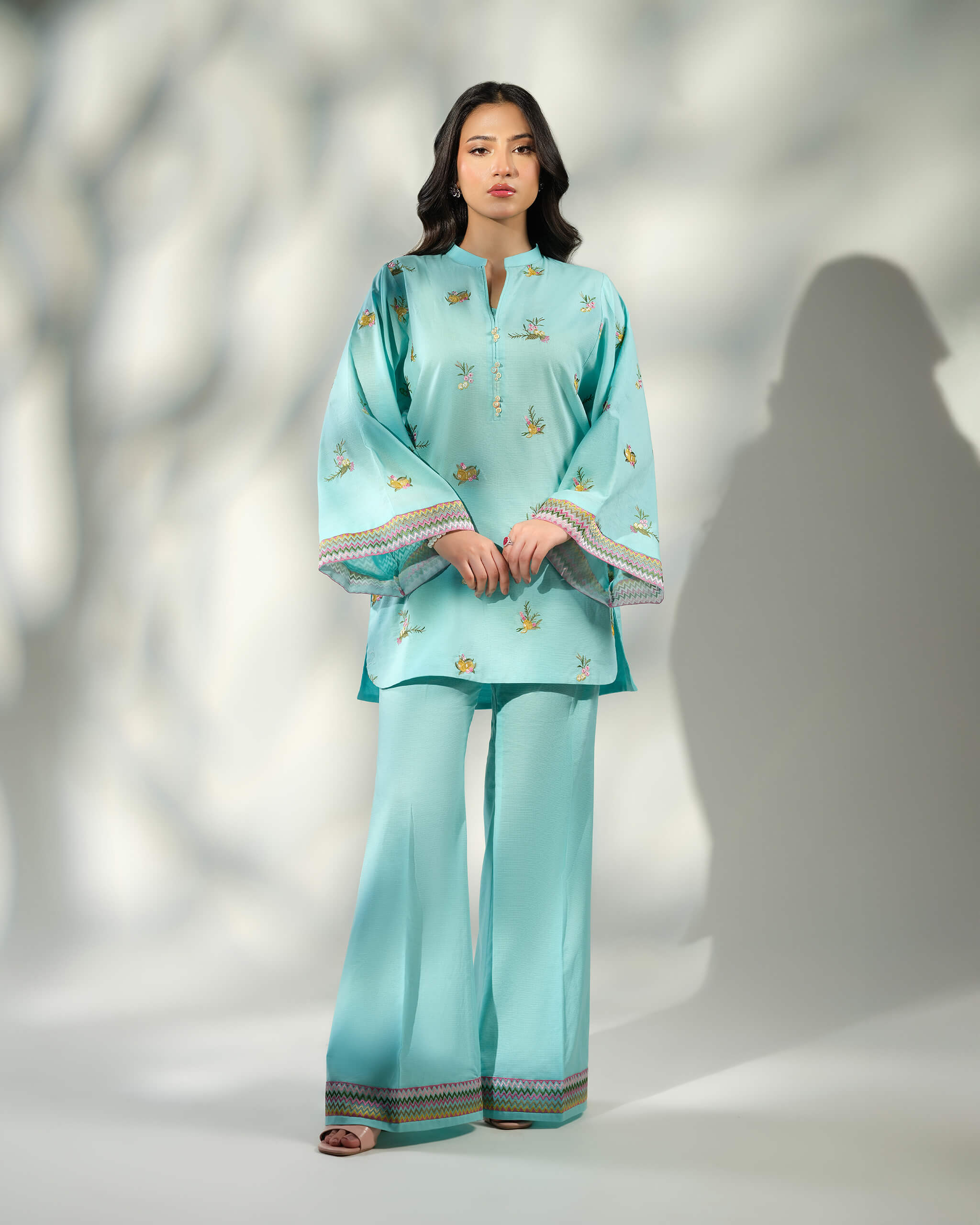 Sinoor Shop pret casual 2025 Amal Light Blue outfit. A woman stands facing forward in a light blue embroidered kurta and flared trousers. The kurta has a band collar and wide, patterned cuffs. The trousers also feature a matching patterned hem. Soft lighting, full view.
