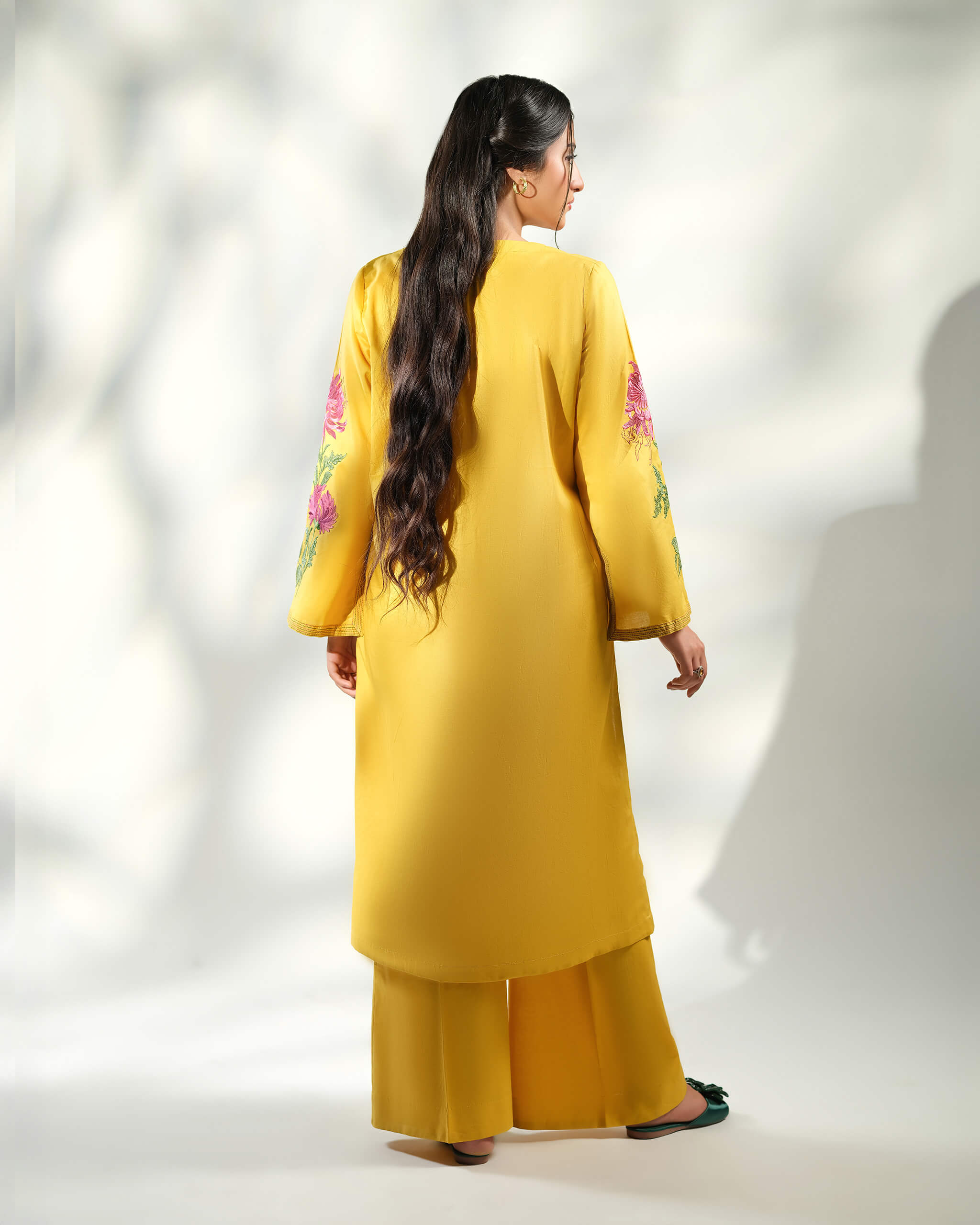 Sinoor Shop pret casual 2025 Arzo Mustard outfit. A woman with long, dark hair is viewed from the back, showcasing the mustard yellow kurta and wide-legged trousers. Floral embroidery accents the sleeves. She is wearing dark green embellished sandals. Back view, studio lighting.