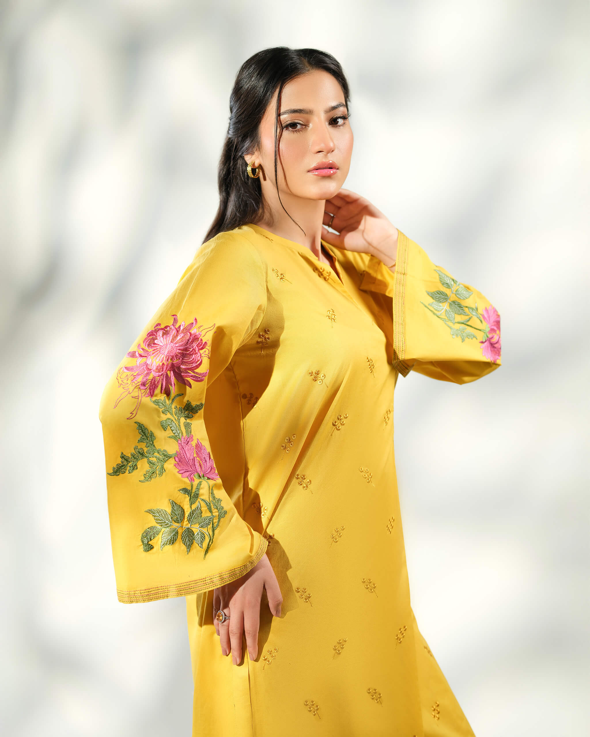 Sinoor Shop pret casual 2025 Arzo Mustard outfit. A woman with dark hair and gold earrings poses in a mustard yellow kurta with vibrant pink and green floral embroidery on the sleeves. She is wearing a ring on her left hand. Close-up shot, studio lighting with shadows.