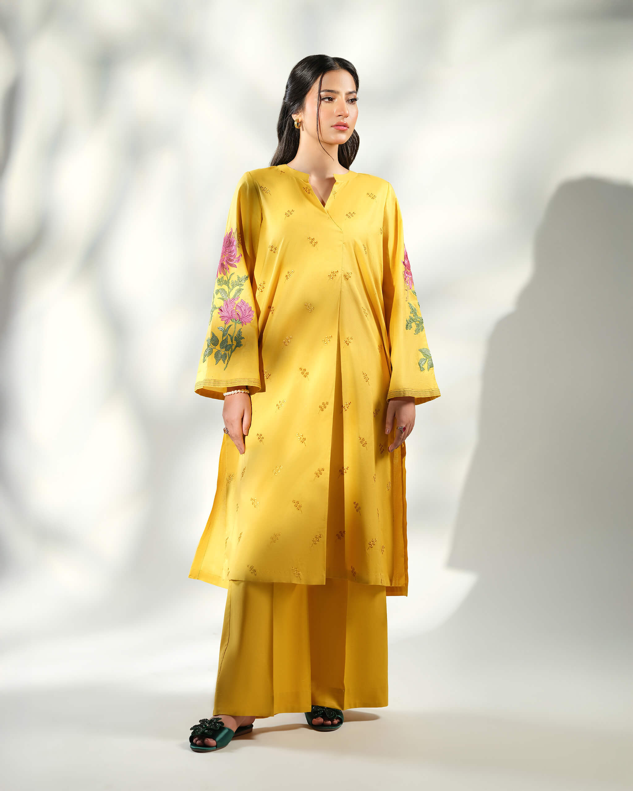 Sinoor Shop pret casual 2025 Arzo Mustard outfit. A woman models a full mustard yellow outfit with a wrap-style kurta featuring floral embroidery on the sleeves and matching wide-legged trousers. She is wearing dark embellished sandals. Full standing shot, studio lighting with shadows.