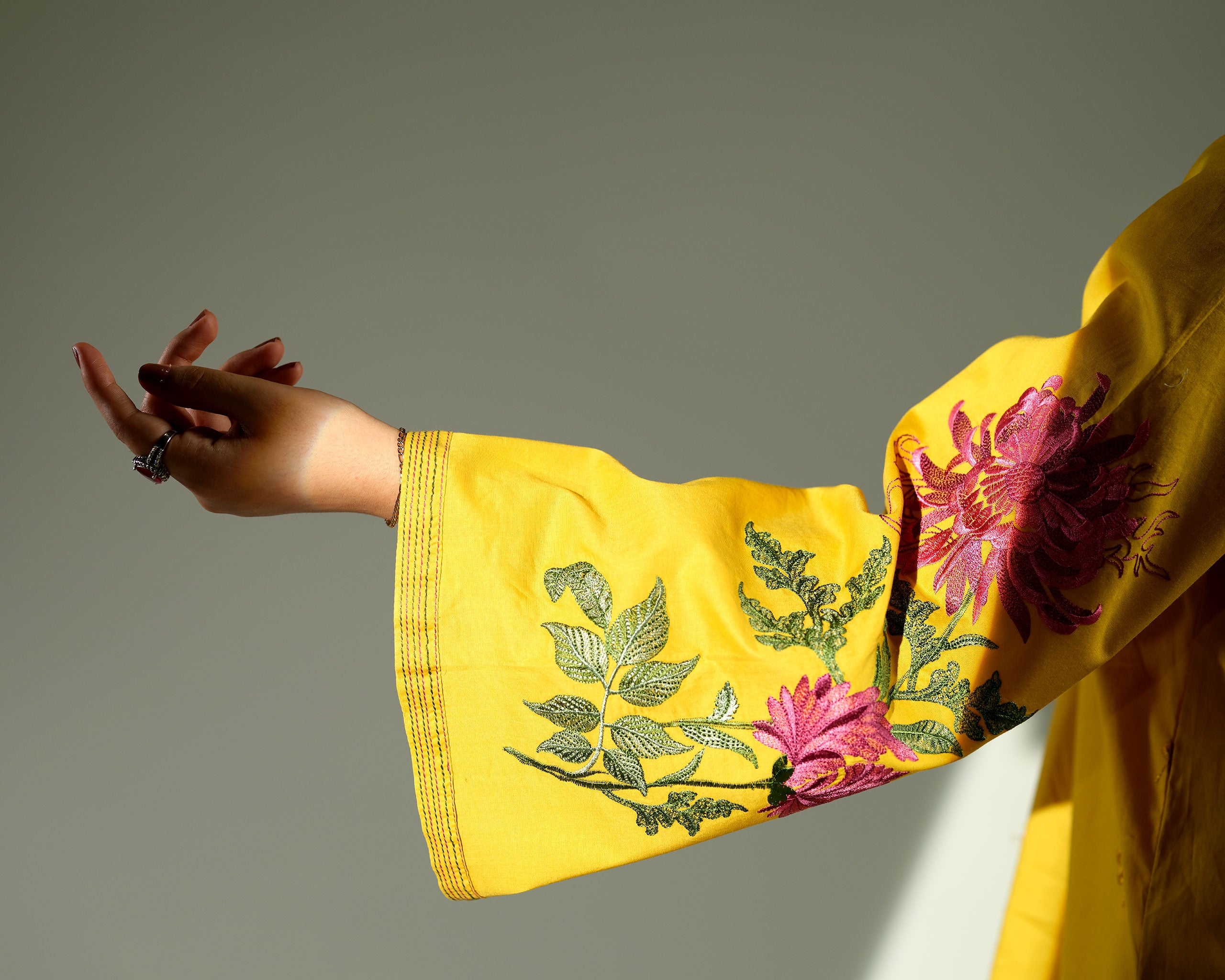 Sinoor Shop pret casual 2025 Arzo Mustard outfit. A woman models a full mustard yellow outfit with a wrap-style kurta featuring floral embroidery on the sleeves and matching wide-legged trousers. She is wearing dark embellished sandals. Full standing shot, studio lighting with shadows.