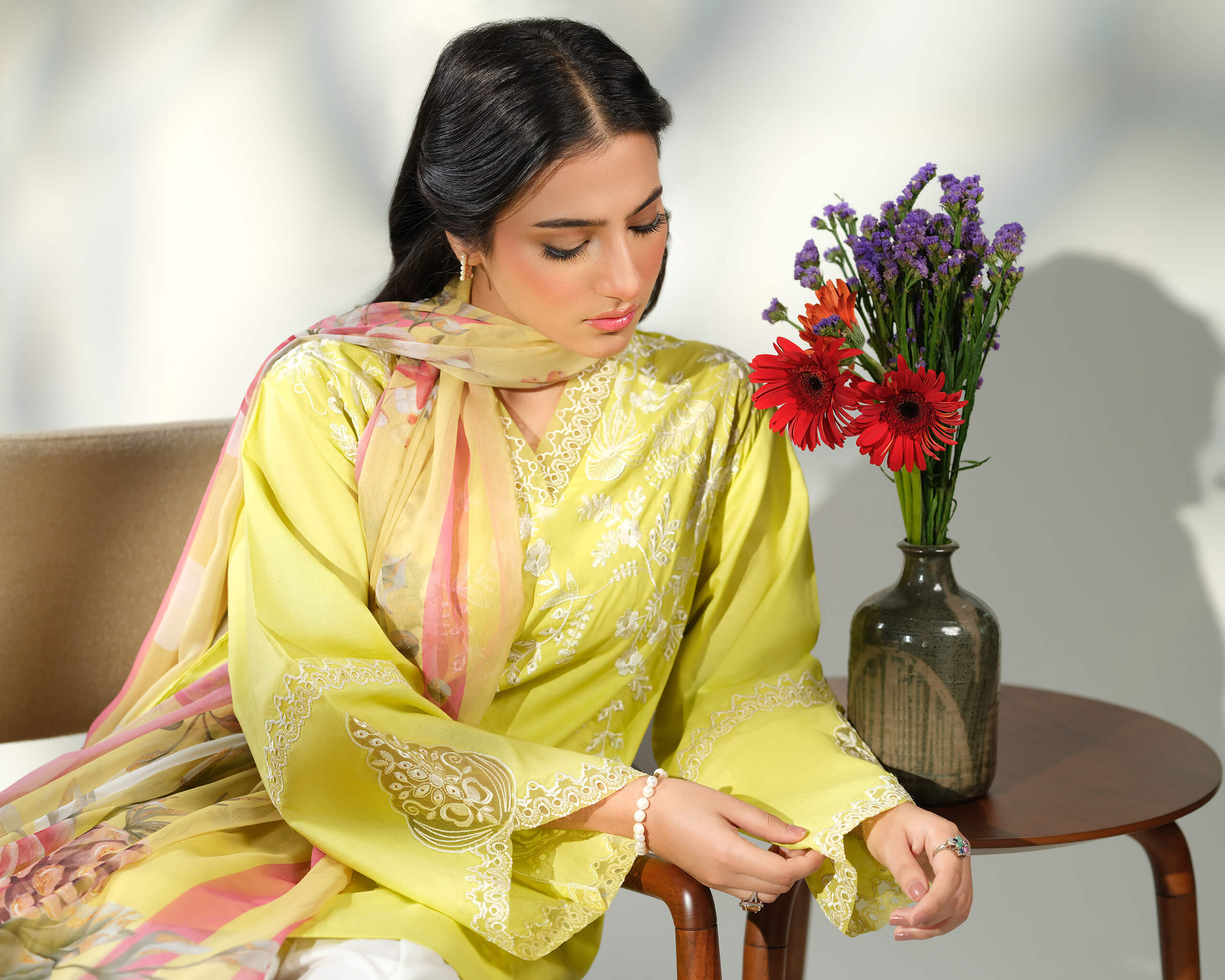 Sinoor Shop pret casual 2025 Asra Lime Yellow outfit. A woman is seated, wearing a lime yellow embroidered kurta with a coordinating dupatta draped around her shoulders. She has dark hair and is wearing delicate jewelry. A vase with red and purple flowers sits on a table beside her. Soft, natural lighting.