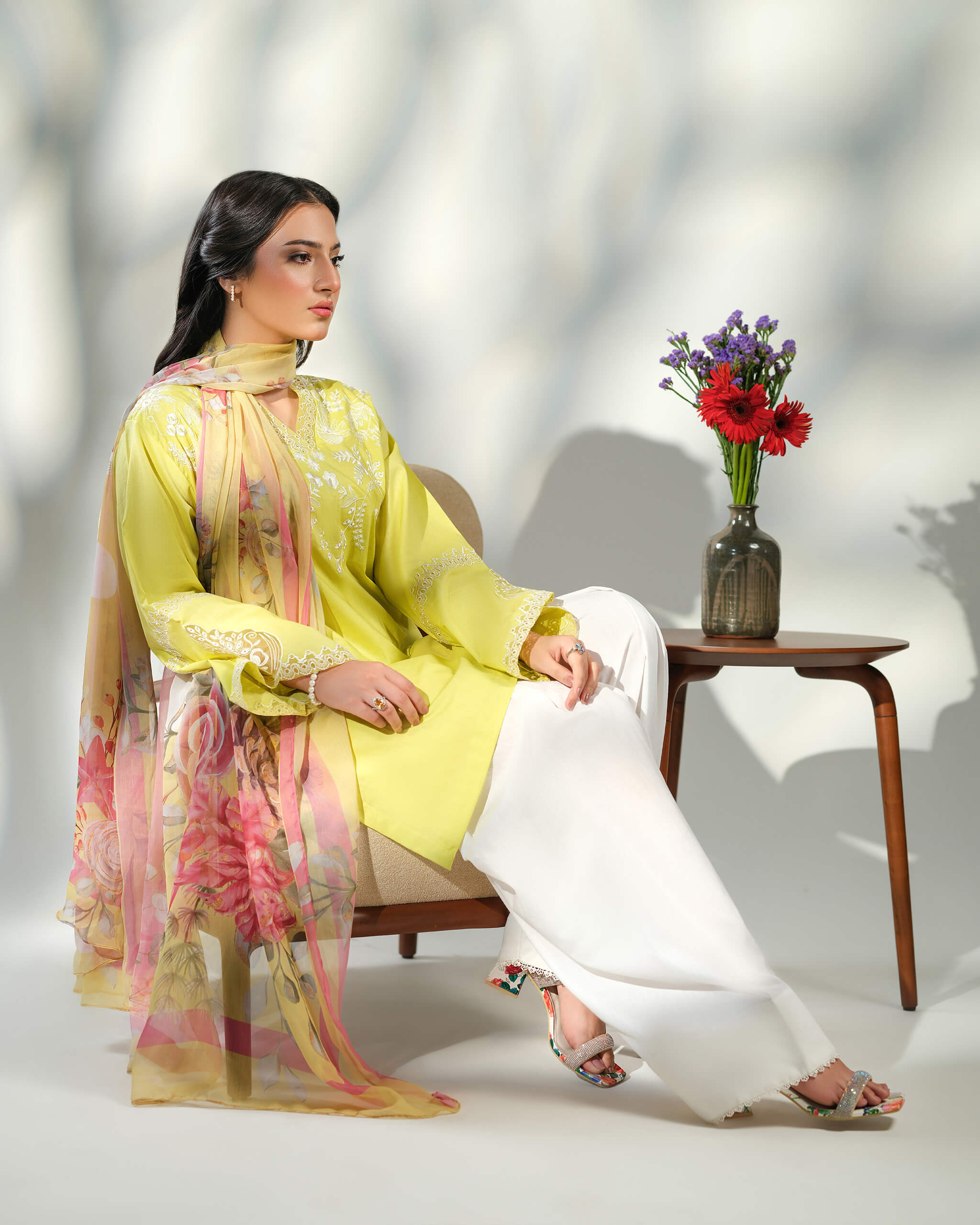 Sinoor Shop pret casual 2025 Asra Lime Yellow outfit. A woman in a lime yellow embroidered kurta and white trousers is seated in a chair. A floral dupatta is draped over her shoulders and lap. A vase of red and purple flowers is on a small table next to her. Soft, studio lighting.