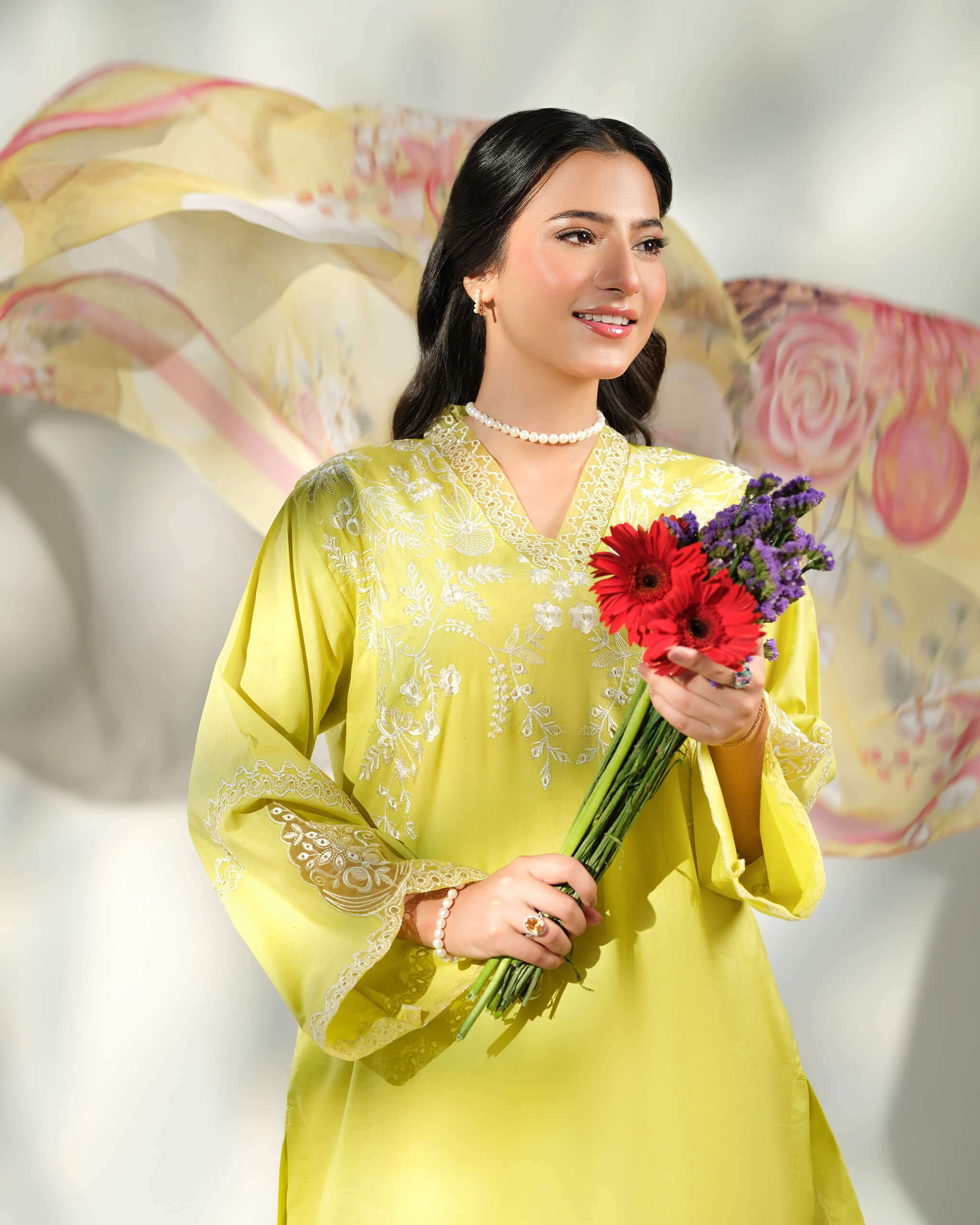 Sinoor Shop pret casual 2025 Asra Lime Yellow outfit. A smiling woman in a lime yellow embroidered kurta and a pearl necklace holds a bouquet of red and purple flowers. A light floral dupatta flows in the background. Close-up, soft studio lighting.
