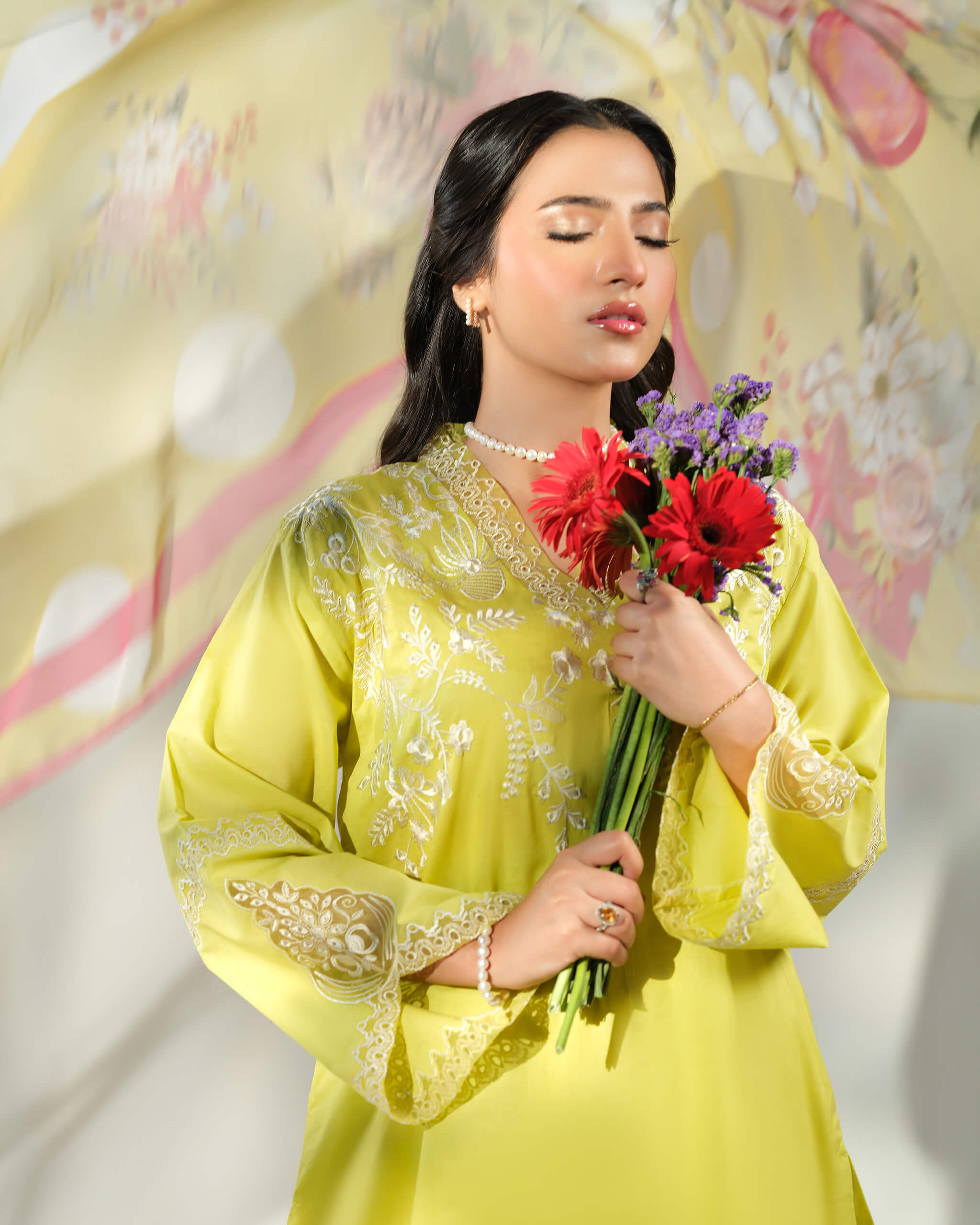 Sinoor Shop pret casual 2025 Asra Lime Yellow outfit. A woman with her eyes closed gently holds a bouquet of red gerbera daisies and purple flowers. She is wearing a lime yellow embroidered kurta and a pearl necklace. A light floral dupatta is behind her. Close-up, soft studio lighting.