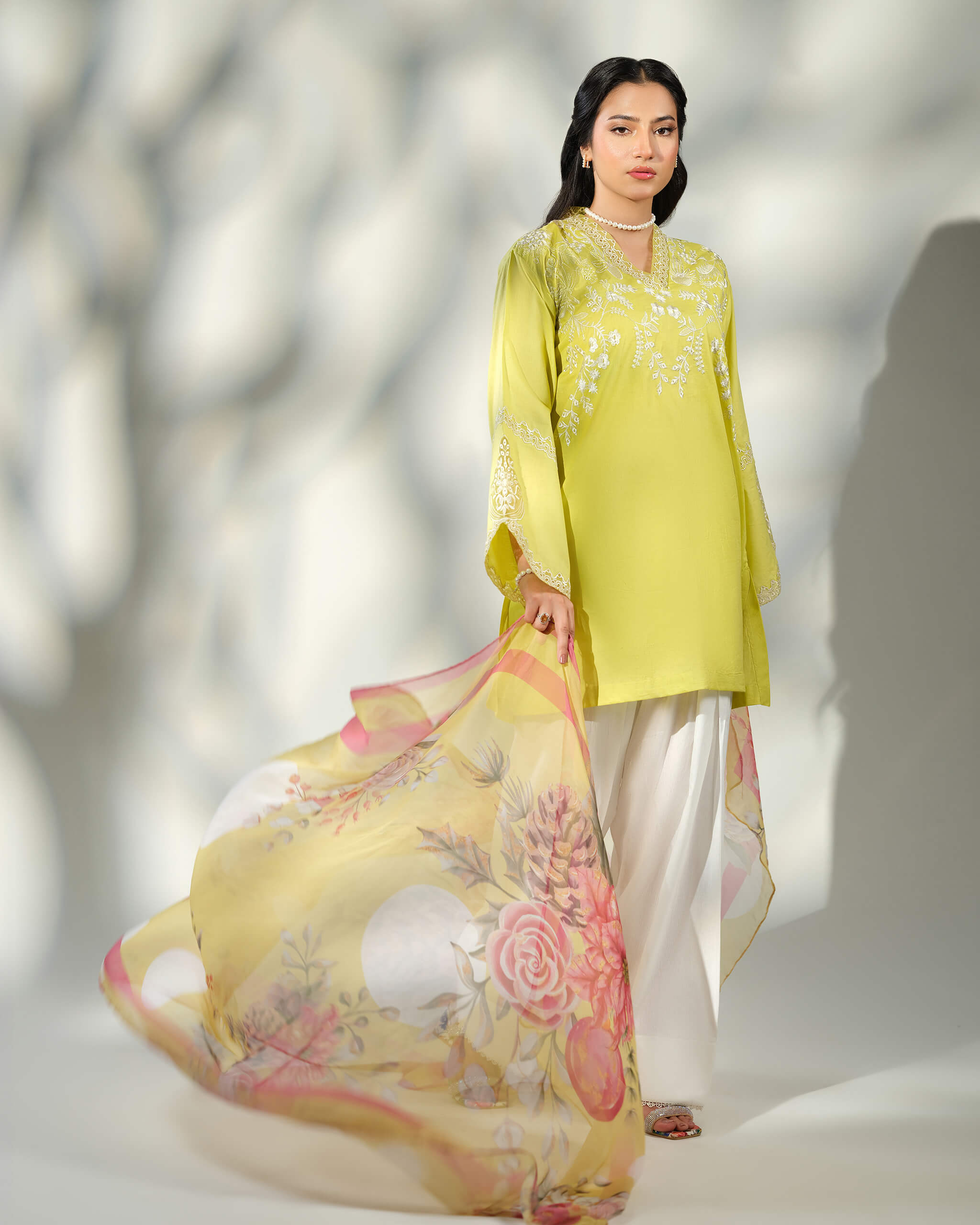 Sinoor Shop pret casual 2025 Asra Lime Yellow outfit. A woman models a lime yellow embroidered kurta with white trousers. She wears a pearl necklace and earrings, and holds a flowing floral dupatta. Full standing shot, soft studio lighting with shadows.