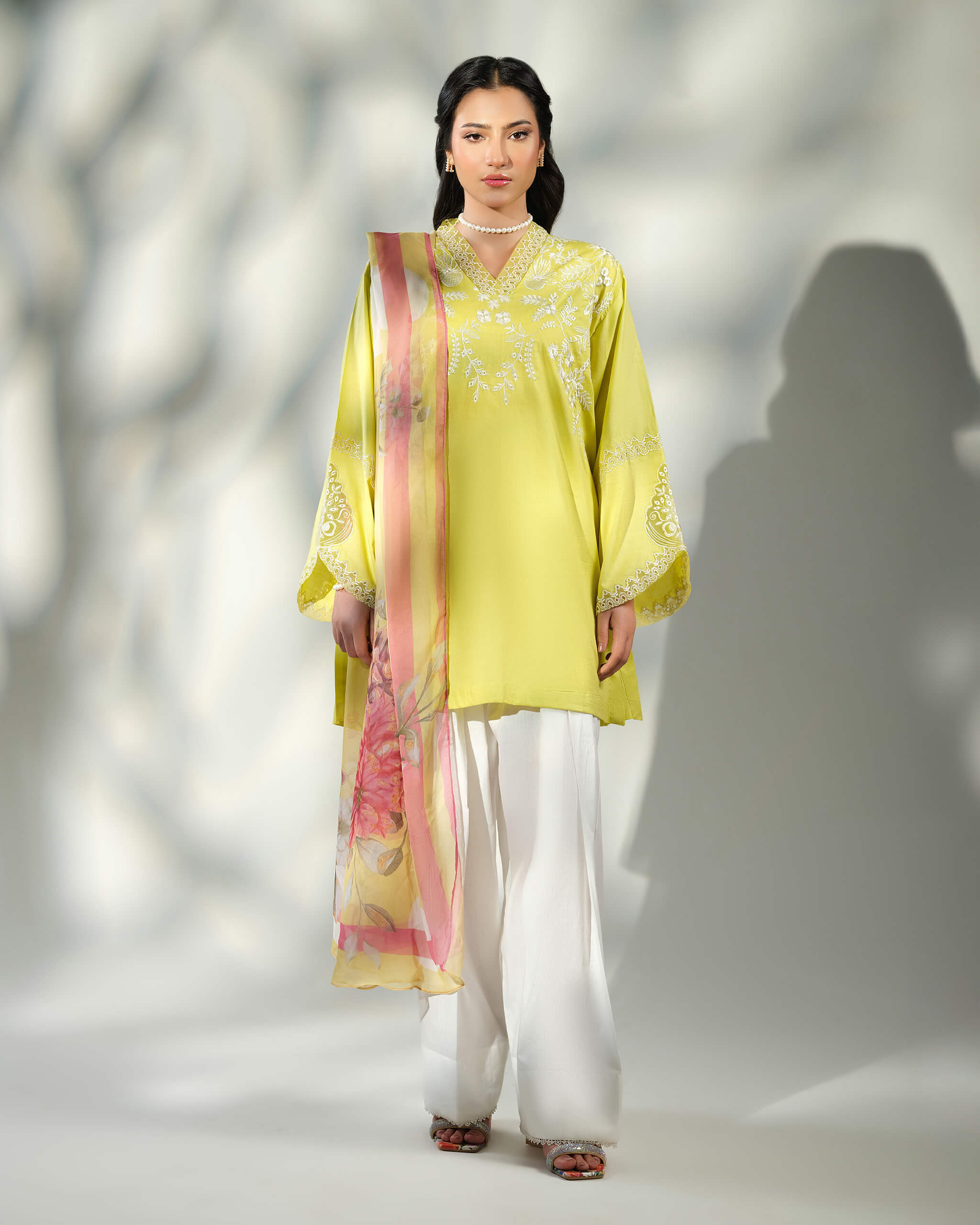 Sinoor Shop pret casual 2025 Asra Lime Yellow outfit. A woman stands in a lime yellow embroidered kurta and white wide-leg trousers. A floral dupatta is draped over one shoulder. She is wearing a pearl necklace and earrings. Full-length shot, studio lighting with shadows.