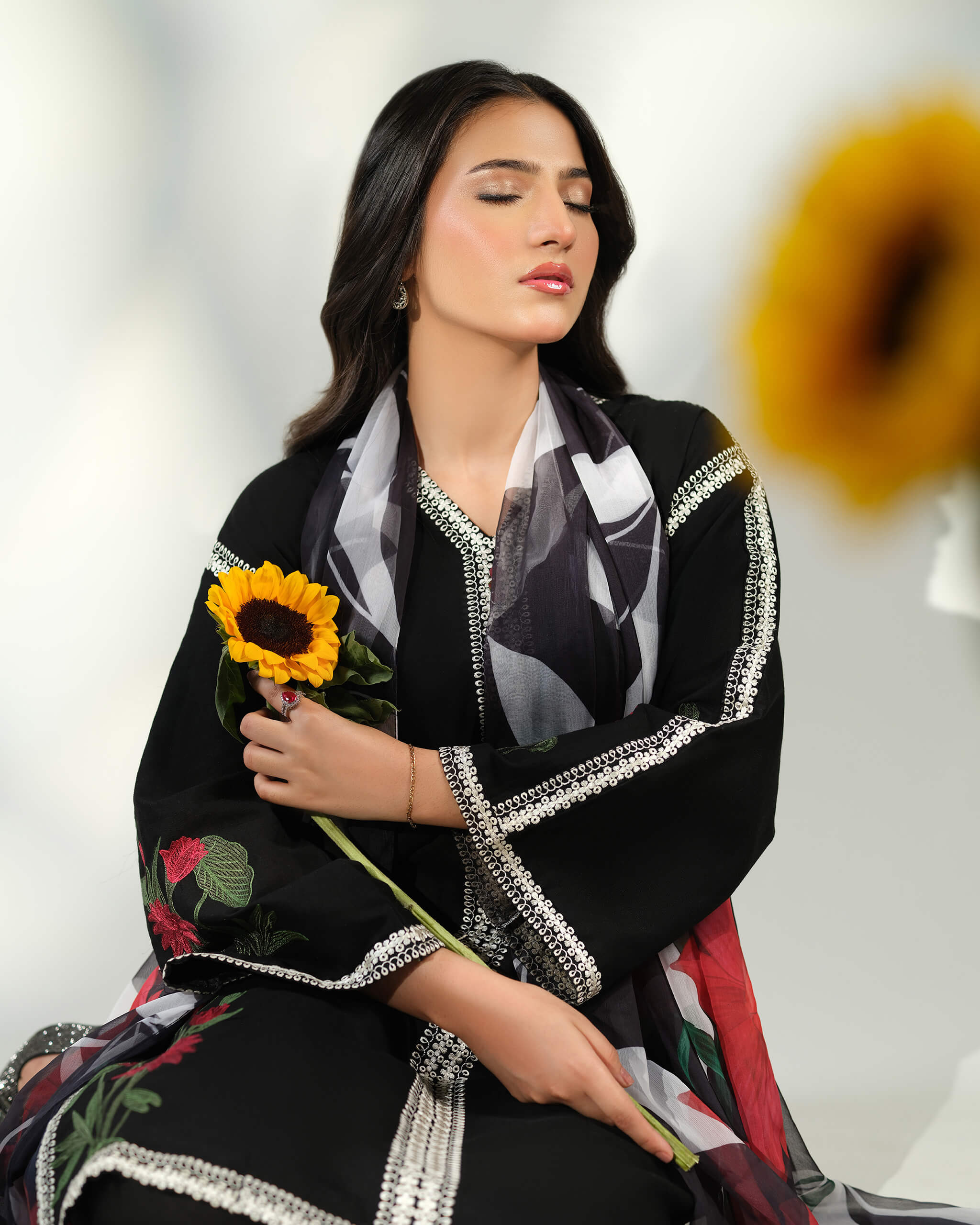 Sinoor Shop pret casual 2025 Ayzel Black outfit. A woman with dark hair and closed eyes holds a sunflower. She is wearing a black embroidered kurta with a printed dupatta draped around her neck. Soft, warm lighting.