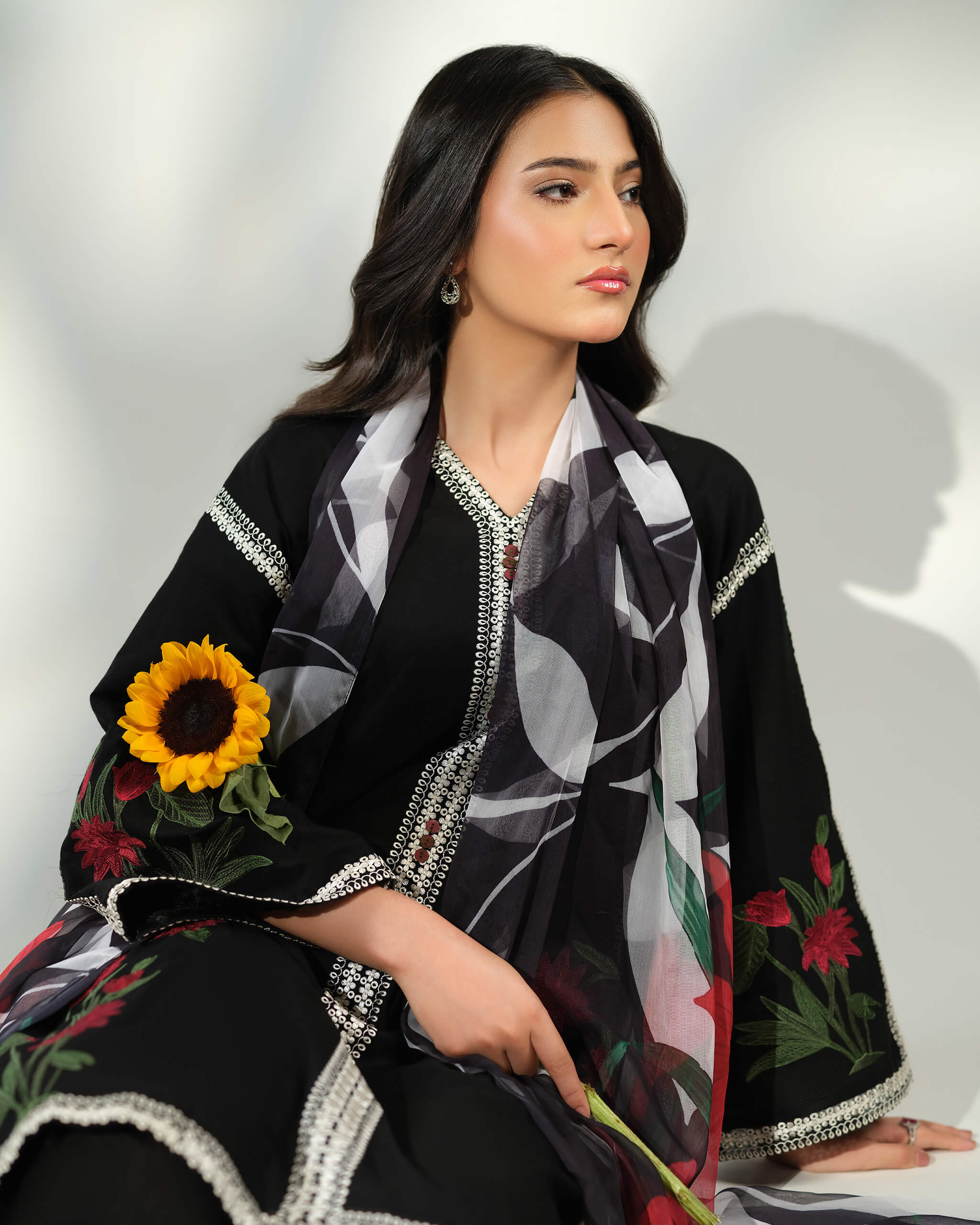 Sinoor Shop pret casual 2025 Ayzel Black outfit. A woman with dark hair looks to the side while holding a sunflower. She is wearing a black kurta with white embroidery details and a floral printed dupatta draped around her shoulders. Soft studio lighting.