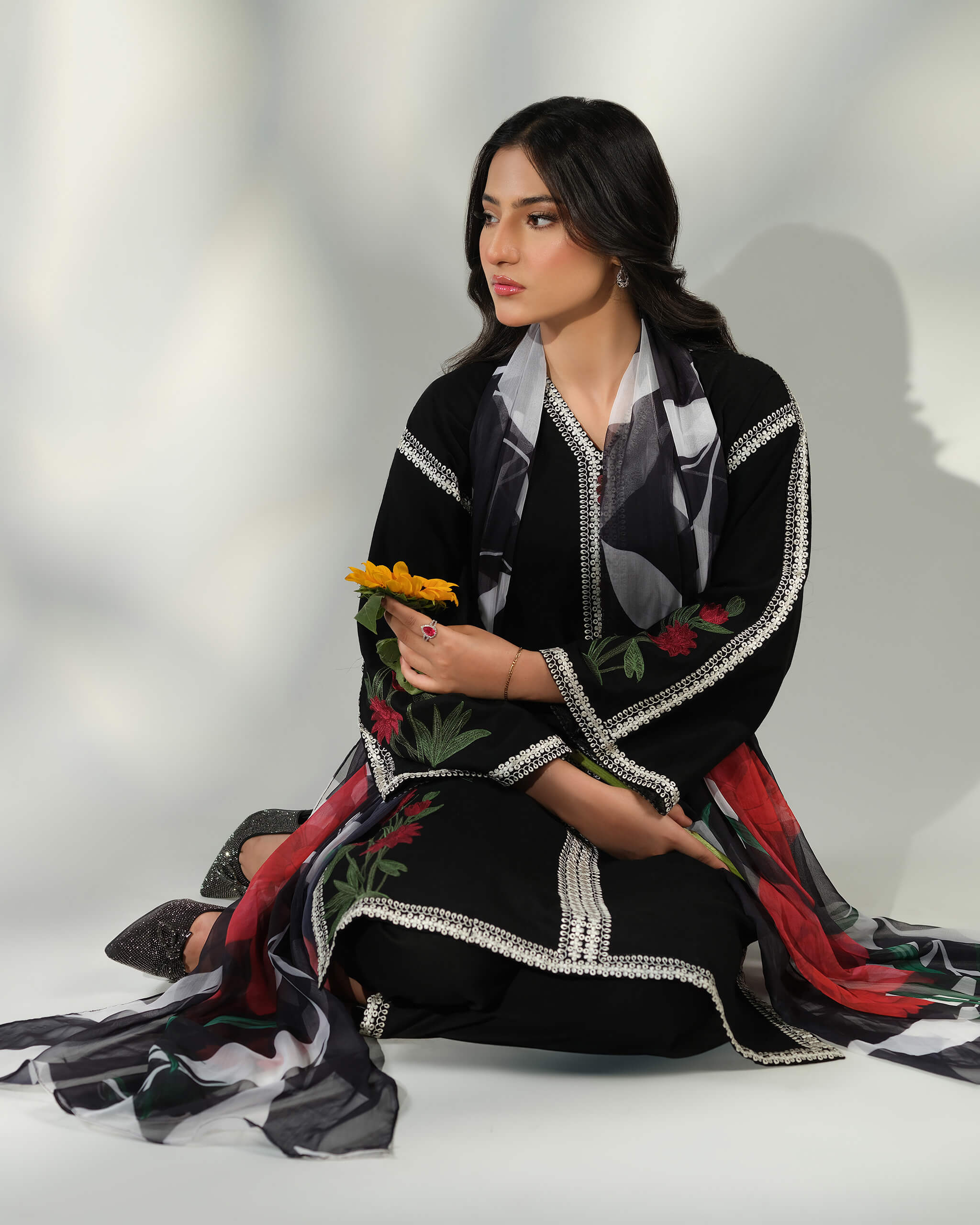 Sinoor Shop pret casual 2025 Ayzel Black outfit. A woman is seated on the floor, holding a small yellow flower. She wears a black embroidered kurta with a digitally printed dupatta. She has dark hair and is wearing dark shoes. Soft studio lighting.