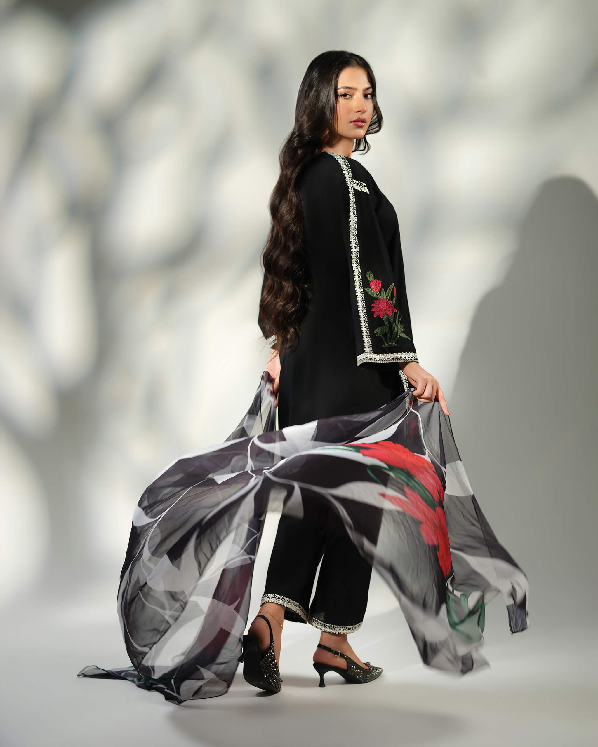 Sinoor Shop pret casual 2025 Ayzel Black outfit. A woman with long, dark hair looks over her shoulder, holding a flowing black and white floral printed dupatta. She is wearing a black embroidered kurta and trousers, and embellished heels. Back view, dynamic studio lighting.