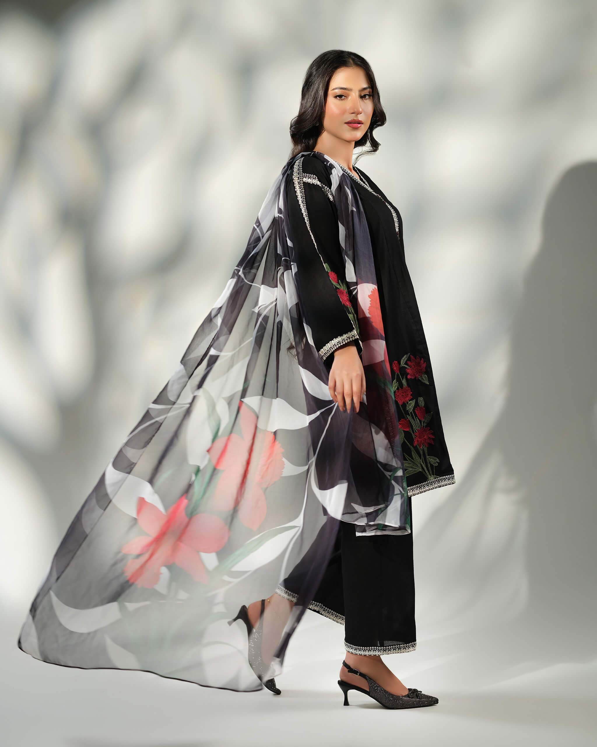 Sinoor Shop pret casual 2025 Ayzel Black outfit. A woman walks, a large black and white floral dupatta flowing behind her. She wears a black embroidered kurta and trousers, paired with embellished heels. Full-length shot, dynamic studio lighting with shadows.