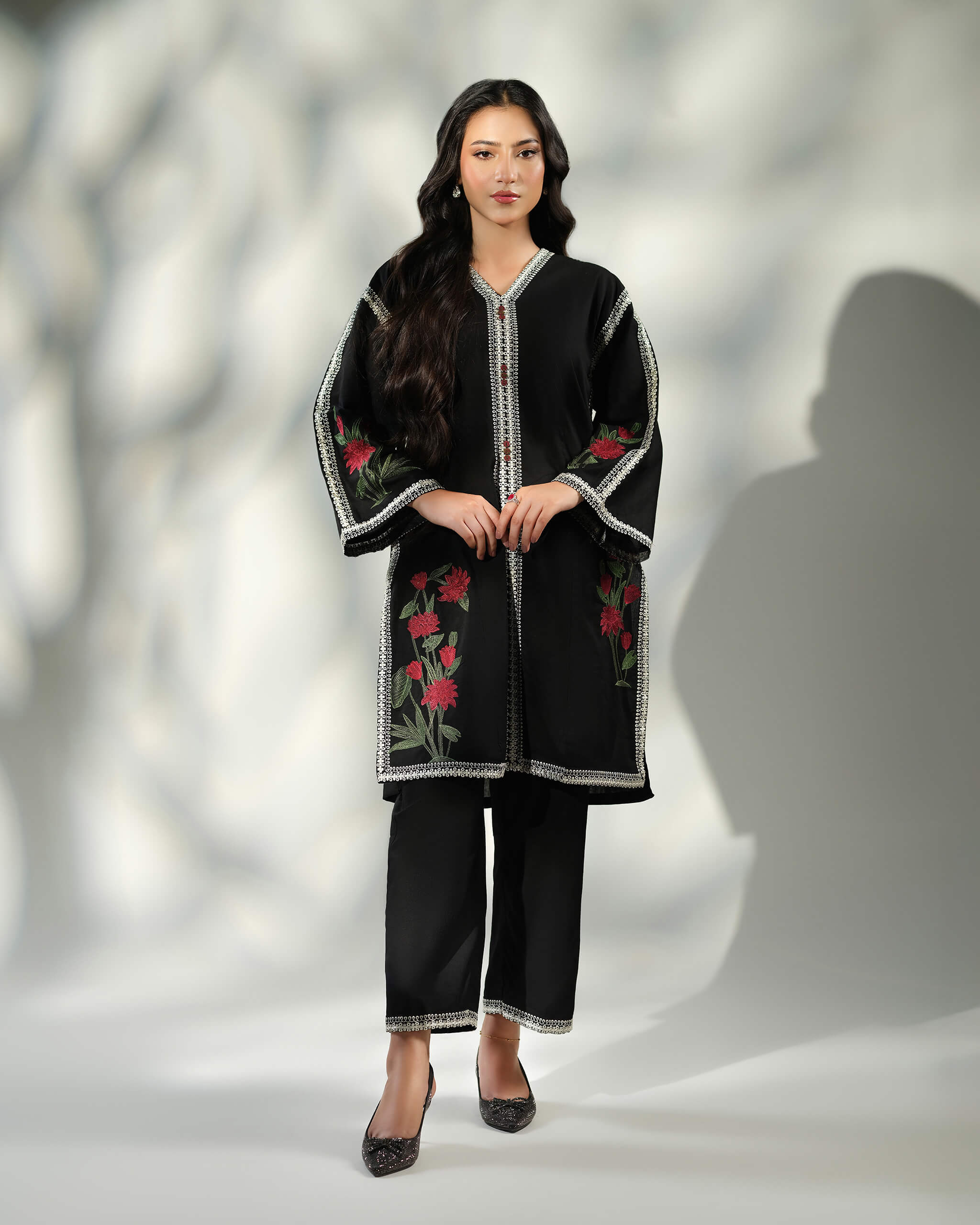 Sinoor Shop pret casual 2025 Ayzel Black outfit. A woman stands facing the camera in a black embroidered kurta and matching trousers. The outfit features detailed white and gold embroidery along the neckline, sleeves, and hem. She is wearing embellished black heels. Full standing shot, studio lighting with shadows.