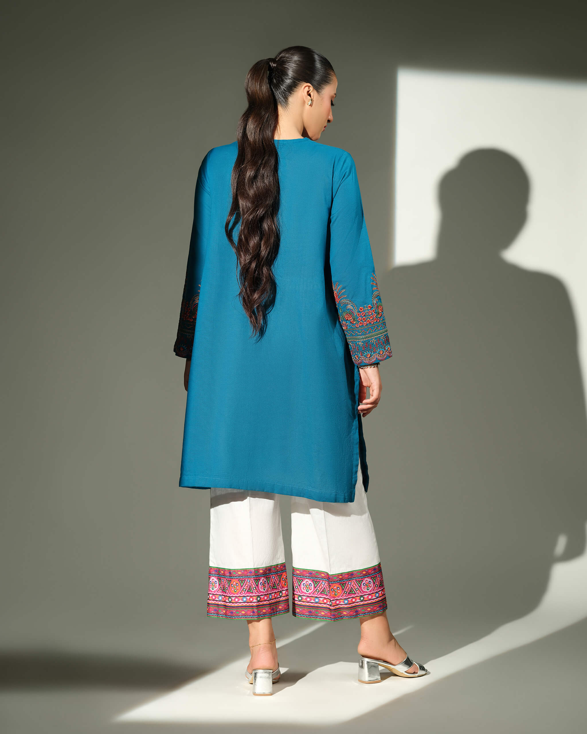 Sinoor Shop pret casual 2025 Mahrosh Teal outfit. A woman with dark hair in a ponytail is seen from the back, wearing a teal kurta and white trousers. The sleeves and the hems of the trousers feature detailed orange and maroon embroidery. She is wearing silver block heels. Back view, studio lighting with shadows.