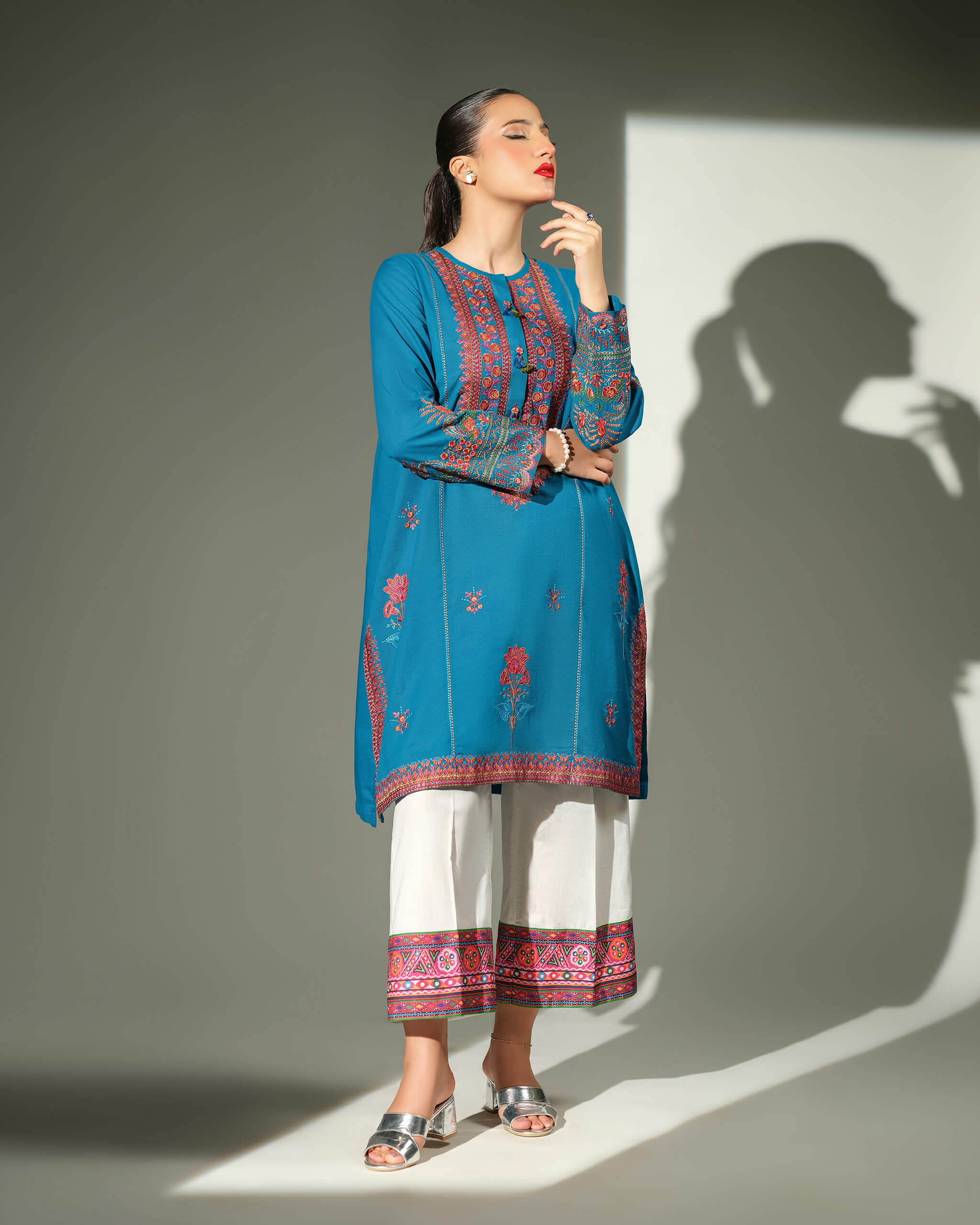 Sinoor Shop pret casual 2025 Mahrosh Teal outfit. A woman stands with her eyes closed, wearing a teal kurta with intricate orange and maroon embroidery and white trousers with a coordinating embroidered hem. She is wearing silver block heels. Full-length shot, studio lighting with a shadow.