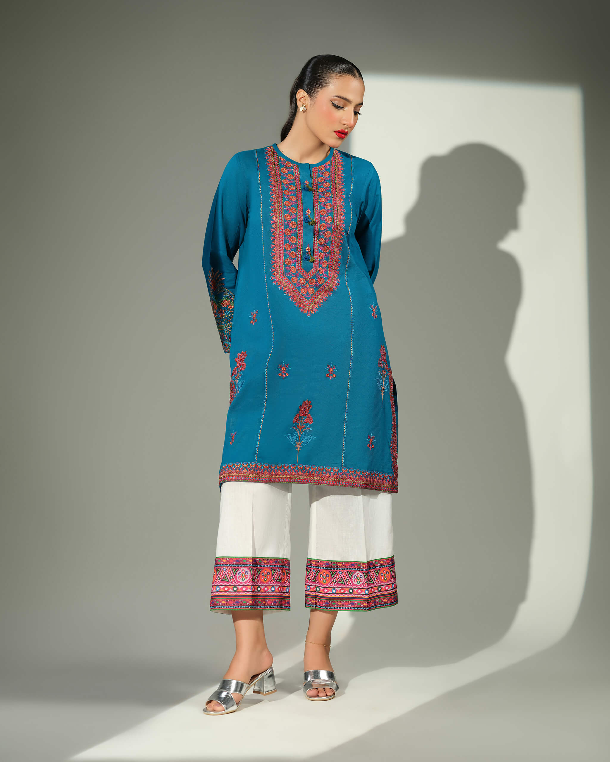 Sinoor Shop pret casual 2025 Mahrosh Teal outfit. A woman models a teal kurta with orange and maroon embroidery and white wide-leg trousers featuring a detailed border. She is wearing silver block heels. Full standing shot, studio lighting with a shadow.