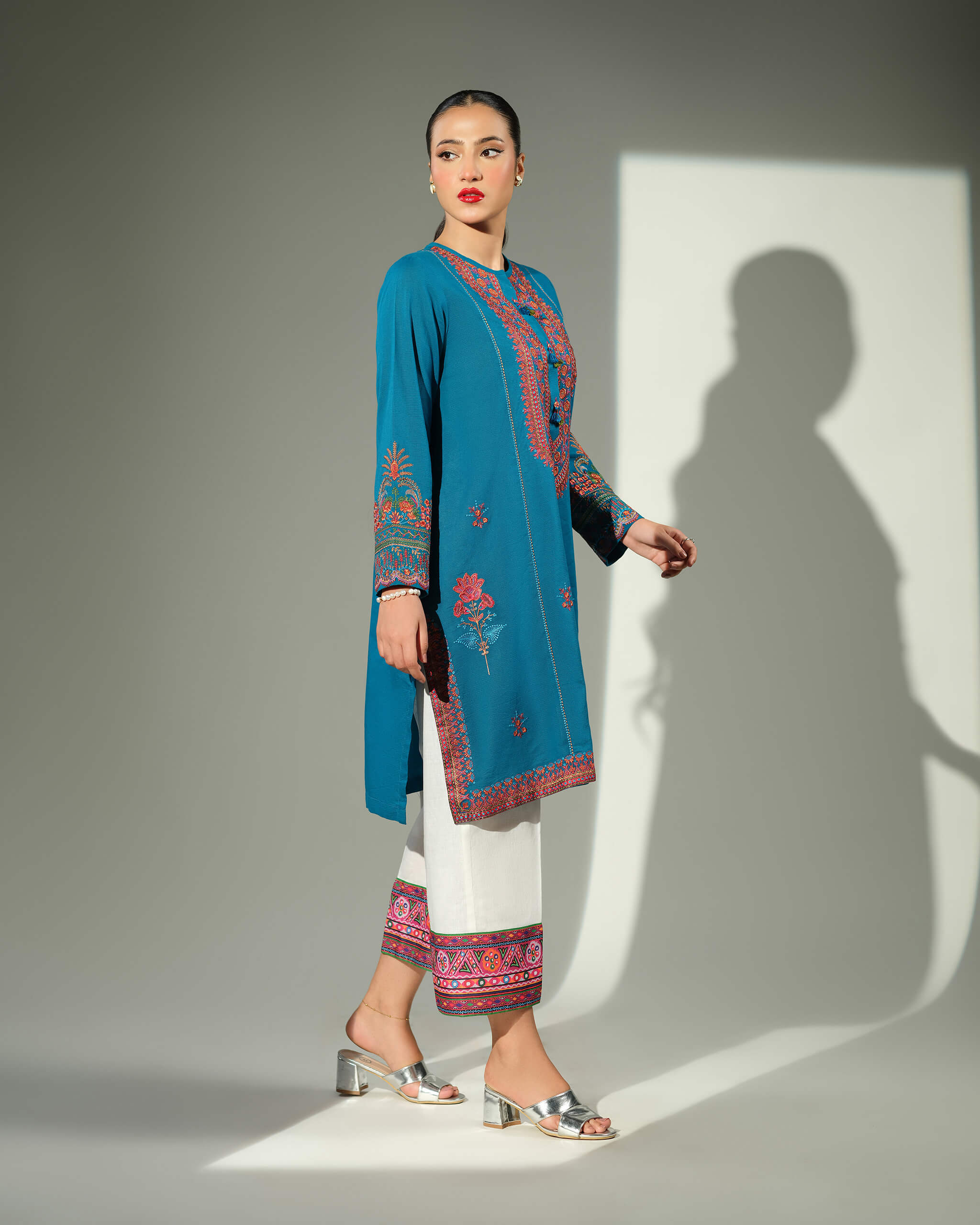 Sinoor Shop pret casual 2025 Mahrosh Teal outfit. A woman walks in profile, wearing a teal kurta with orange and maroon embroidery and white trousers with an embroidered hem. She is wearing silver block heels. Full-length shot, studio lighting with a strong shadow.