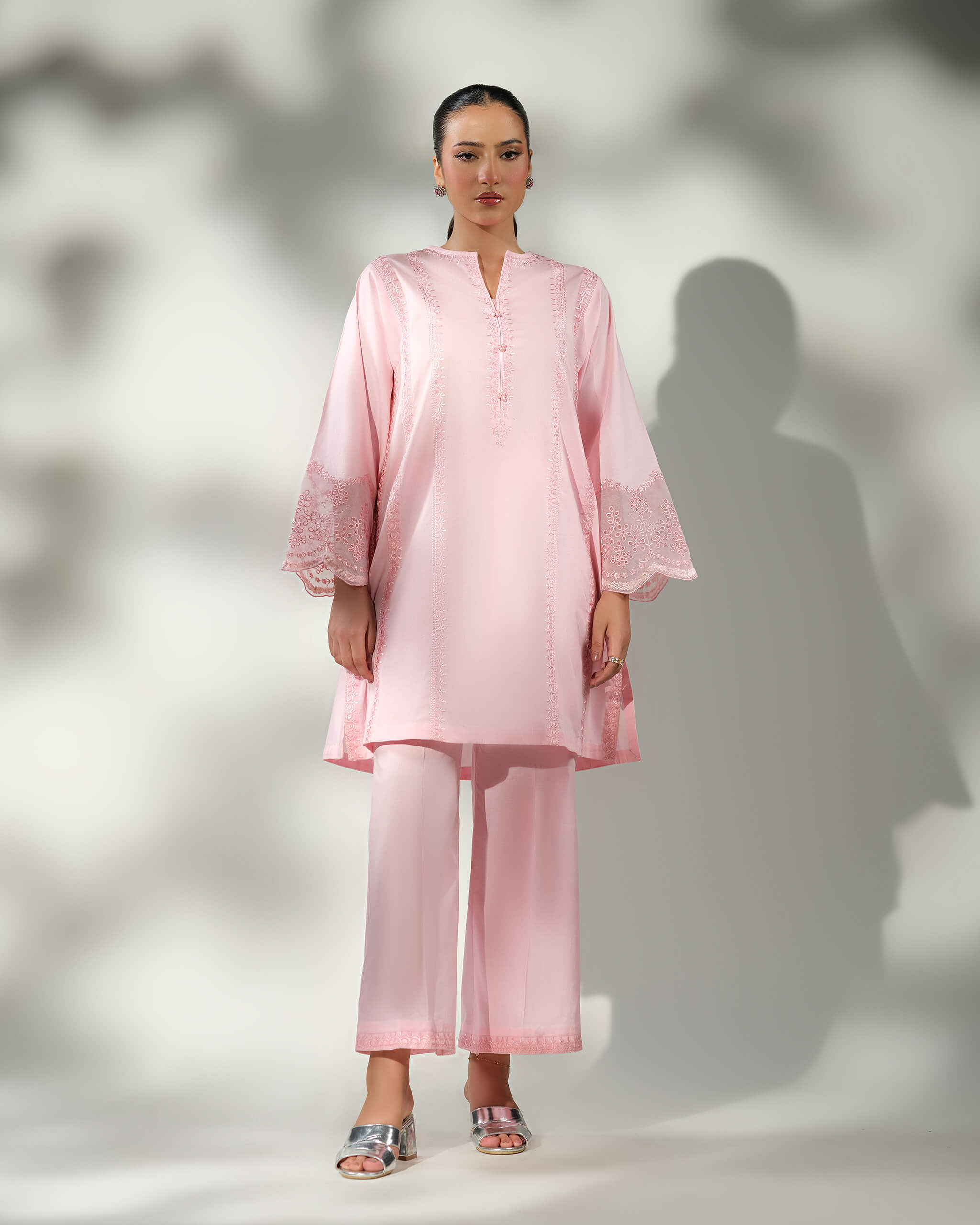Sinoor Shop pret casual 2025 Mira Baby Pink outfit. A woman stands facing forward in a light baby pink kurta and wide-leg trousers. The outfit features delicate white embroidery on the sleeves and front panel. She is wearing silver block heels. Full standing shot, soft studio lighting with shadows.
