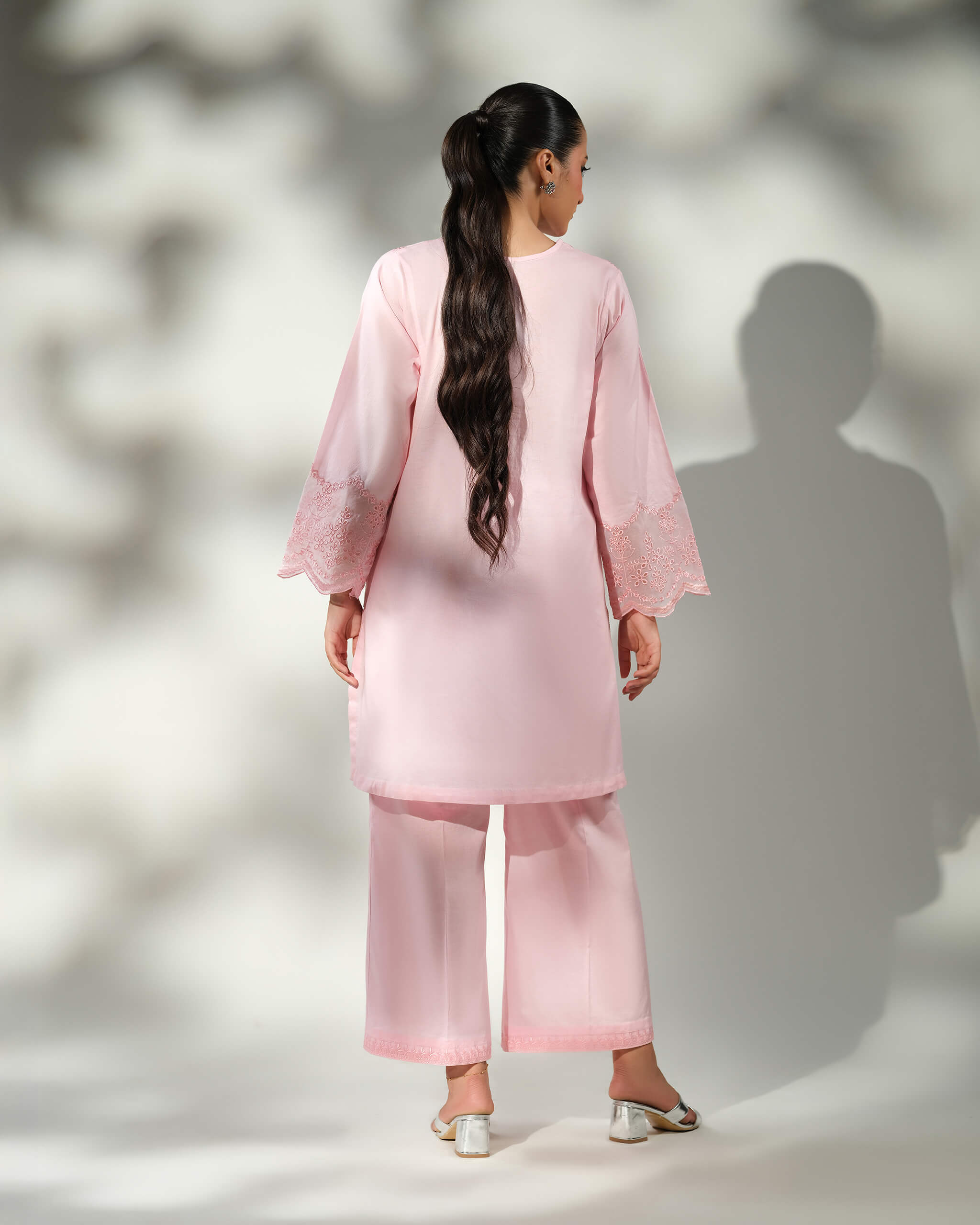 Sinoor Shop pret casual 2025 Mira Baby Pink outfit. A woman with long, dark hair in a ponytail is viewed from the back, modeling a light baby pink kurta and wide-leg trousers. The sleeves feature delicate white embroidery. She is wearing silver block heels. Back view, soft studio lighting with shadows.