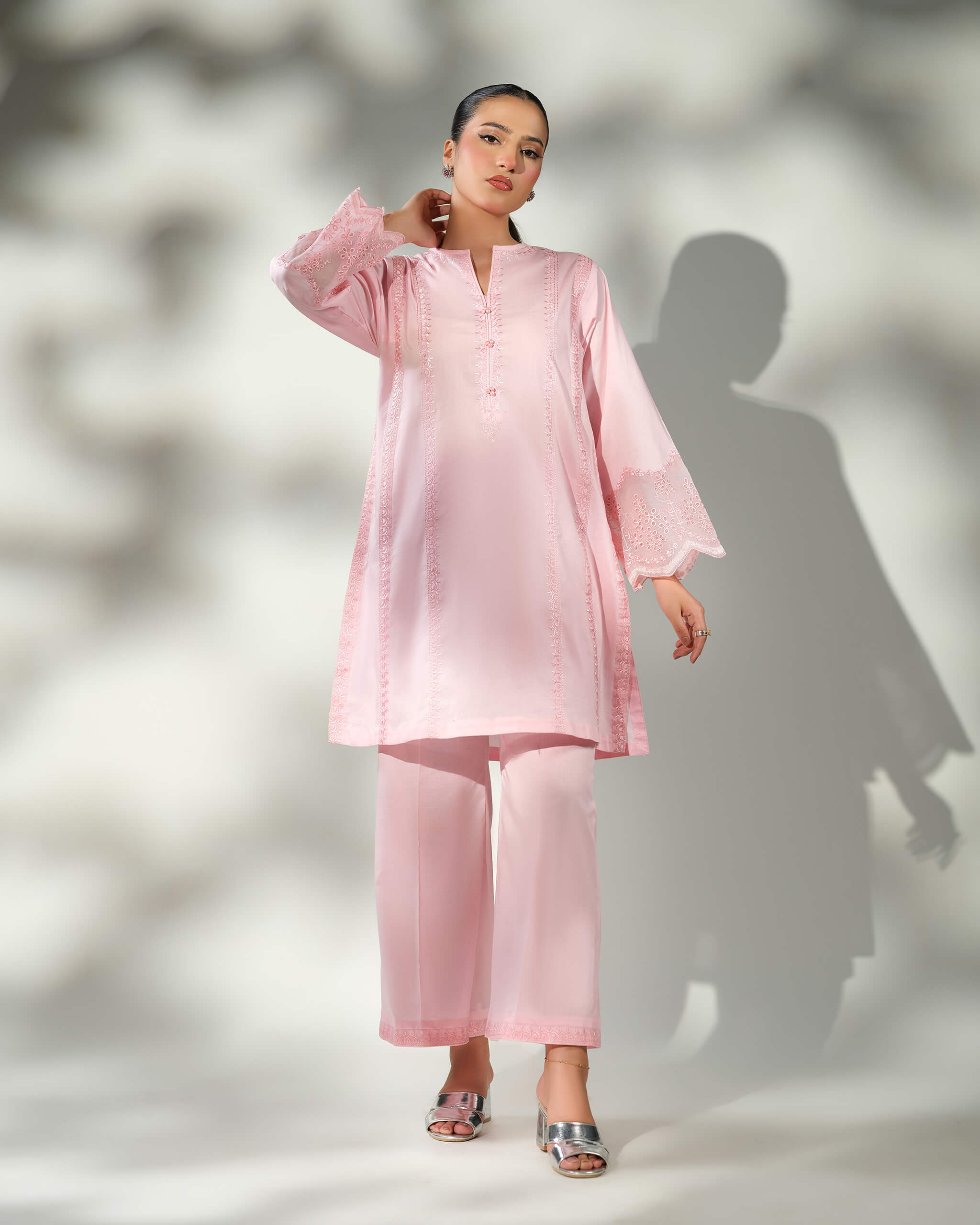 Sinoor Shop pret casual 2025 Mira Baby Pink outfit. A woman stands with one hand touching her hair, wearing a light baby pink kurta and wide-leg trousers. The sleeves of the kurta have intricate white embroidery. She is wearing silver block heels. Full-length shot, soft studio lighting with a shadow.