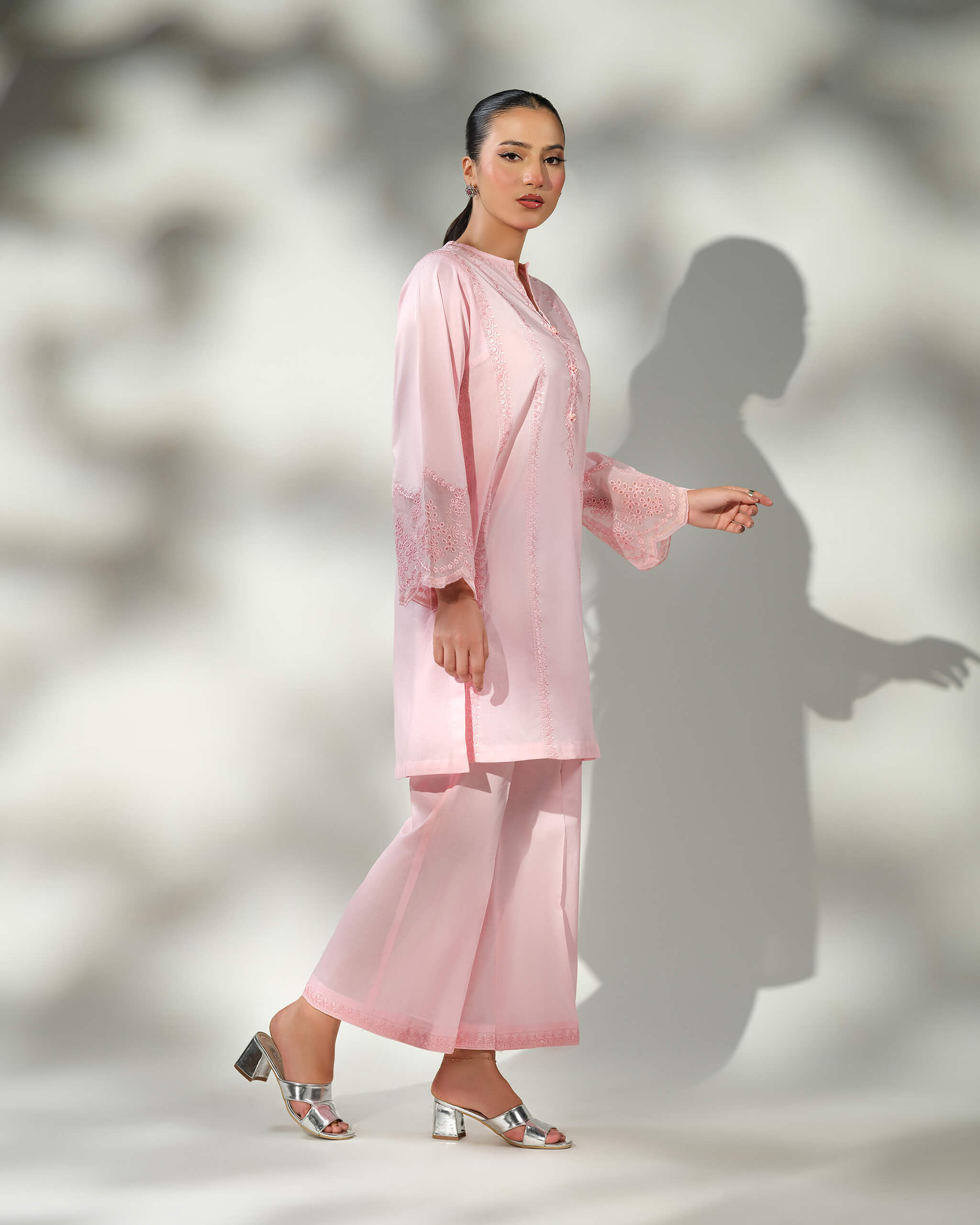 Sinoor Shop pret casual 2025 Mira Baby Pink outfit. A woman walks in profile, wearing a light baby pink kurta with delicate white embroidery on the sleeves and matching wide-leg trousers. She is wearing silver block heels. Full-length shot, soft studio lighting with a strong shadow.