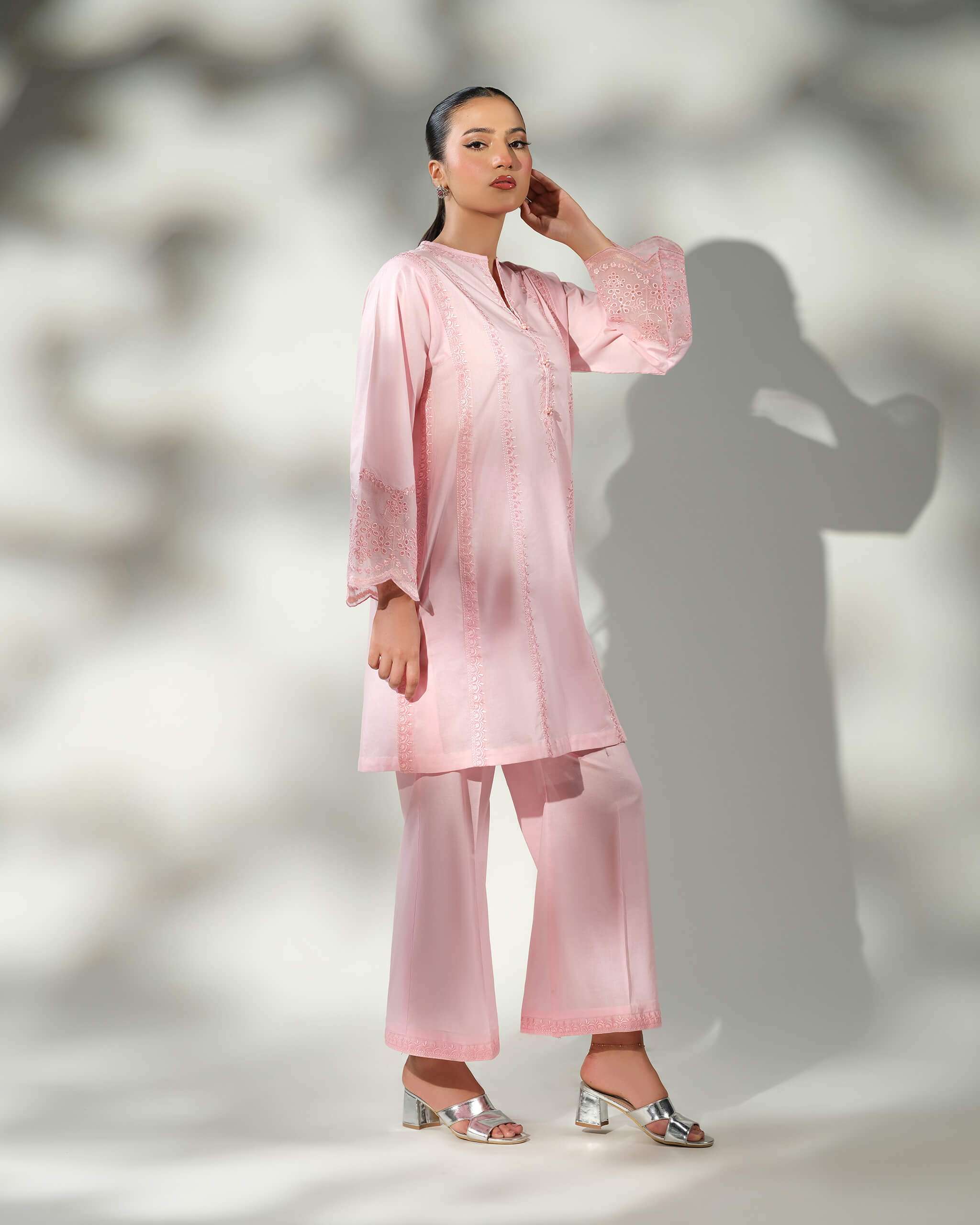 Sinoor Shop pret casual 2025 Mira Baby Pink outfit. A woman poses with one hand on her neck, wearing a light baby pink kurta with subtle white embroidery and matching wide-leg trousers. She is wearing silver block heels. Full standing shot, soft studio lighting with a shadow.