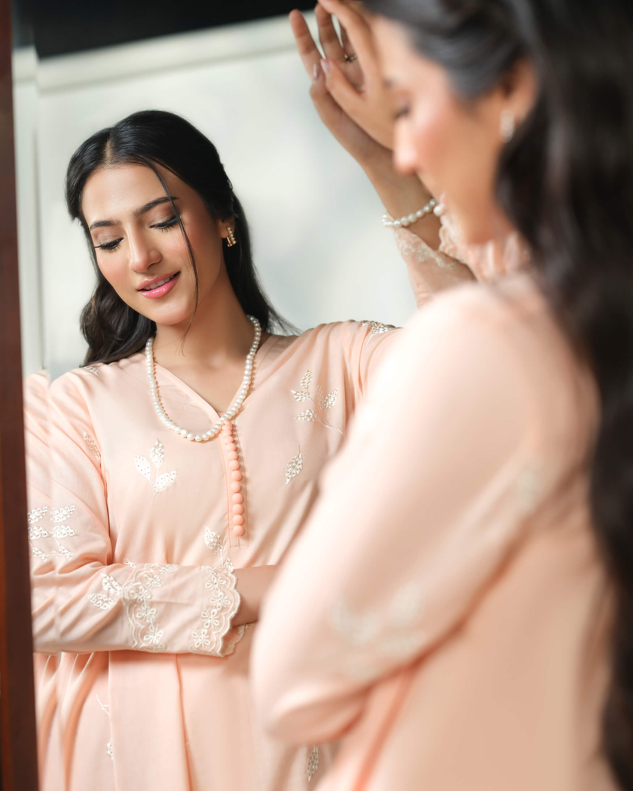 Sinoor Shop pret casual 2025 Naz Peach outfit. A woman with dark hair looks at her reflection in a mirror. She is wearing a light peach-colored kurta with delicate white embroidery and a long pearl necklace. Soft, natural indoor lighting.