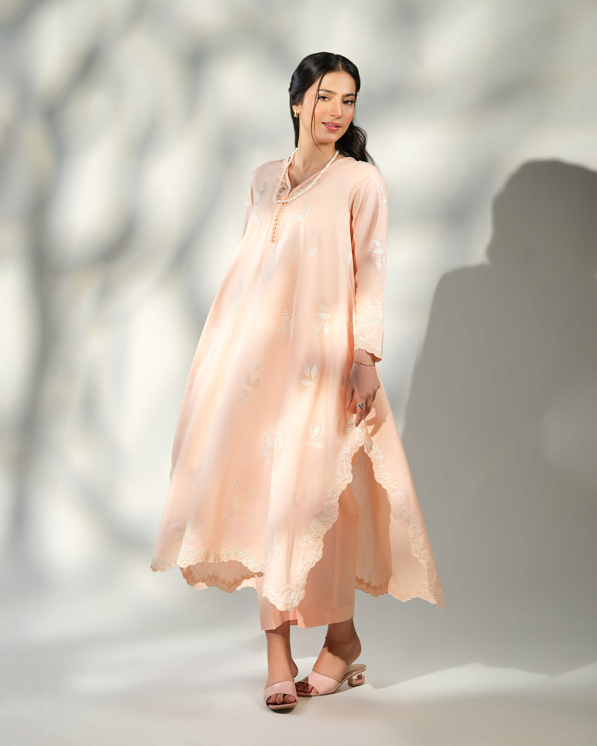 Sinoor Shop pret casual 2025 Naz Peach outfit. A woman stands smiling in a flowy, light peach-colored kurta with white embroidery and matching trousers. She is wearing a long pearl necklace and light-colored heels. Full-length shot, soft studio lighting with shadows.