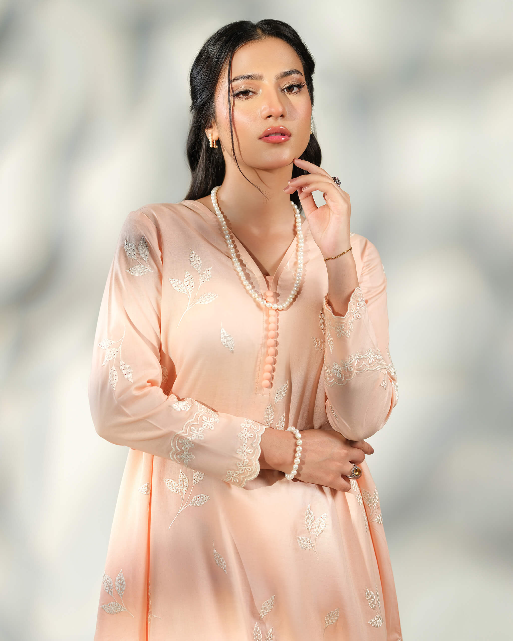 Sinoor Shop pret casual 2025 Naz Peach outfit. A woman looks at the camera, wearing a light peach-colored kurta with delicate white embroidery. She has dark hair and is adorned with a long pearl necklace, a pearl bracelet, and rings. Close-up shot, soft studio lighting with shadows.