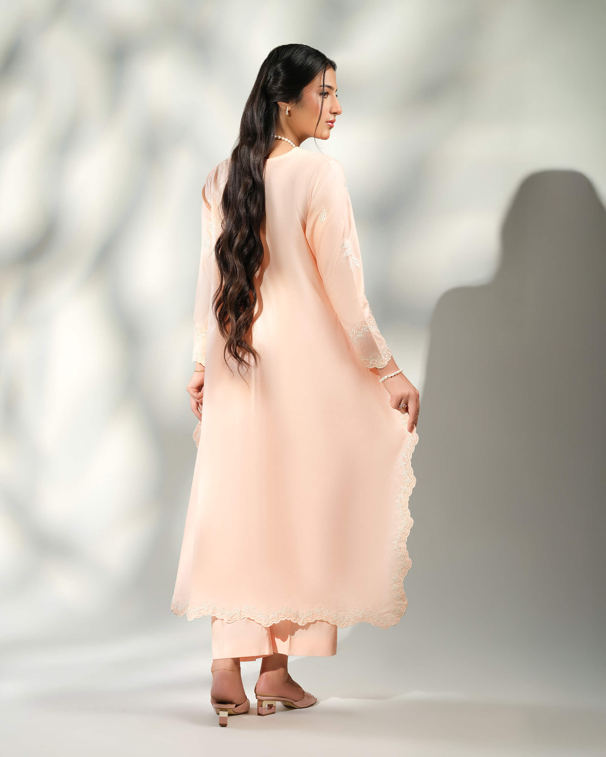 Sinoor Shop pret casual 2025 Naz Peach outfit. A woman with long, dark hair is seen from the back, modeling a flowy, light peach-colored kurta with white embroidery and matching trousers. She is wearing a long pearl necklace and light-colored heels. Back view, soft studio lighting with a shadow.
