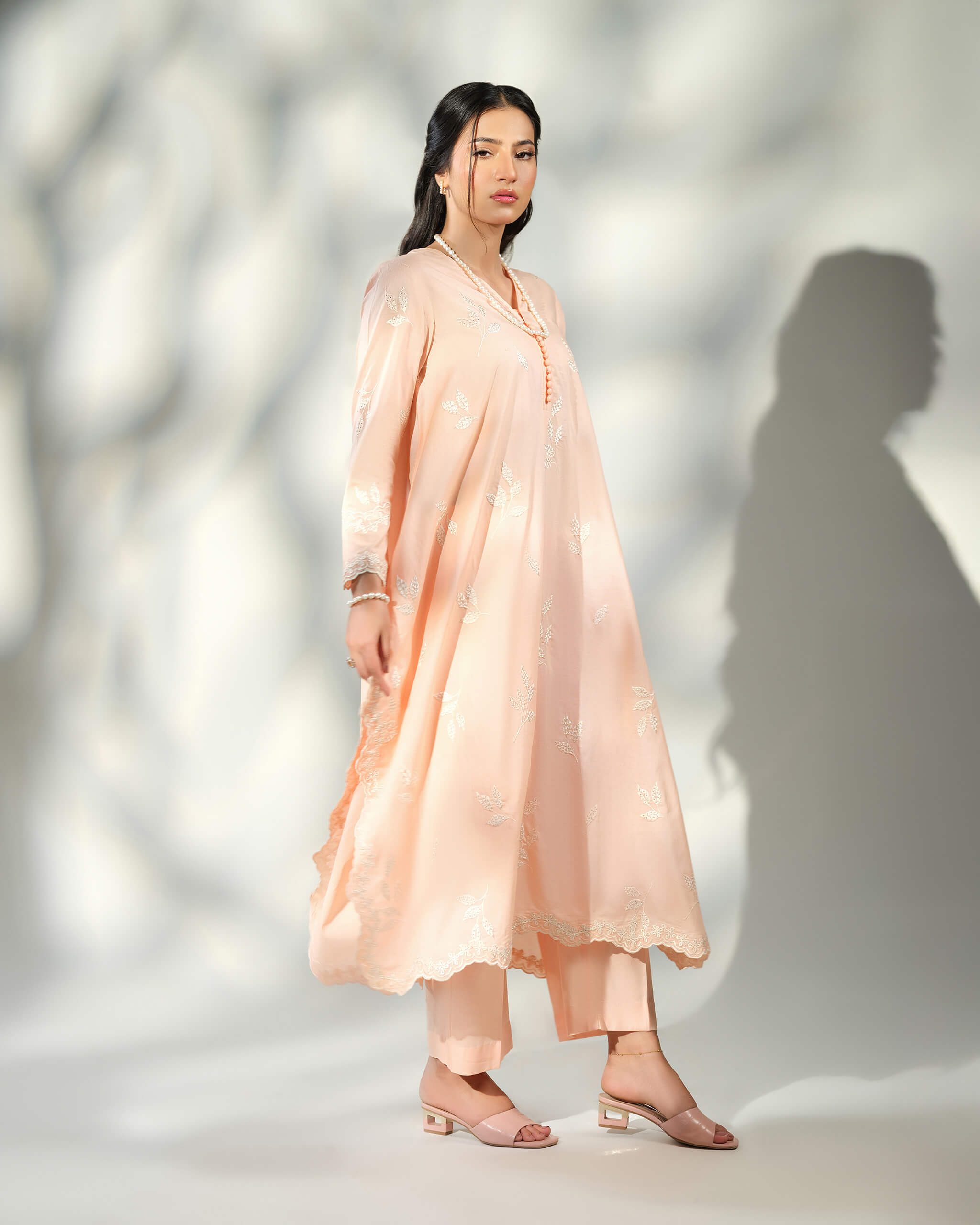 Sinoor Shop pret casual 2025 Naz Peach outfit. A woman stands in a light peach-colored, flowy kurta with white embroidery and matching straight trousers. She is wearing a long pearl necklace and light pink heels. Full-length shot, soft studio lighting with a shadow.