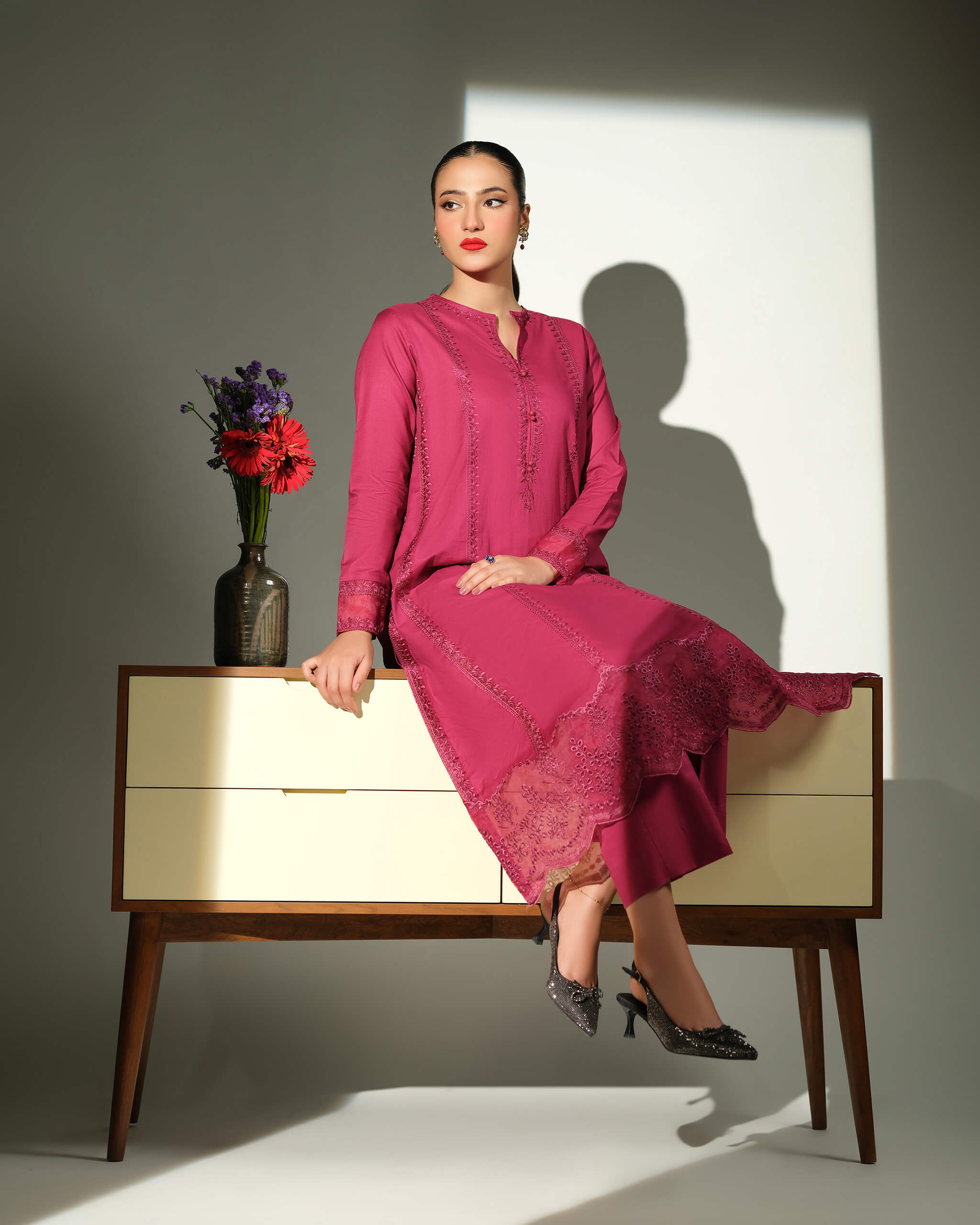 Sinoor Shop pret casual 2025 Sible Dark Pink outfit. A woman with red lipstick and dark earrings sits on a mid-century modern cabinet, wearing a dark pink embroidered kurta and trousers. A vase of red and purple flowers is on the cabinet. Studio lighting with a defined shadow.