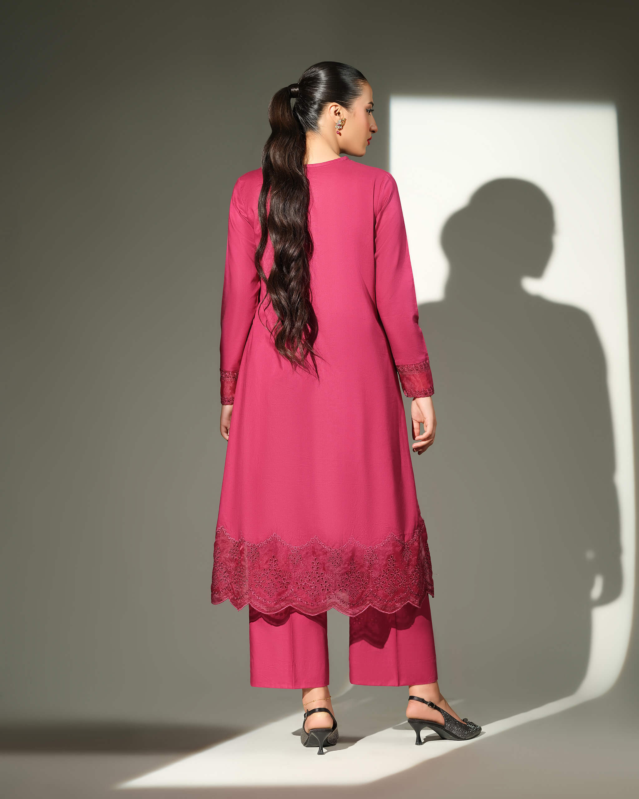 Sinoor Shop pret casual 2025 Sible Dark Pink outfit. A woman with long, dark hair in a ponytail is viewed from the back, wearing a dark pink kurta with embroidered details on the hem and sleeves, and matching trousers. She is wearing embellished black heels. Back view, studio lighting with a strong shadow.