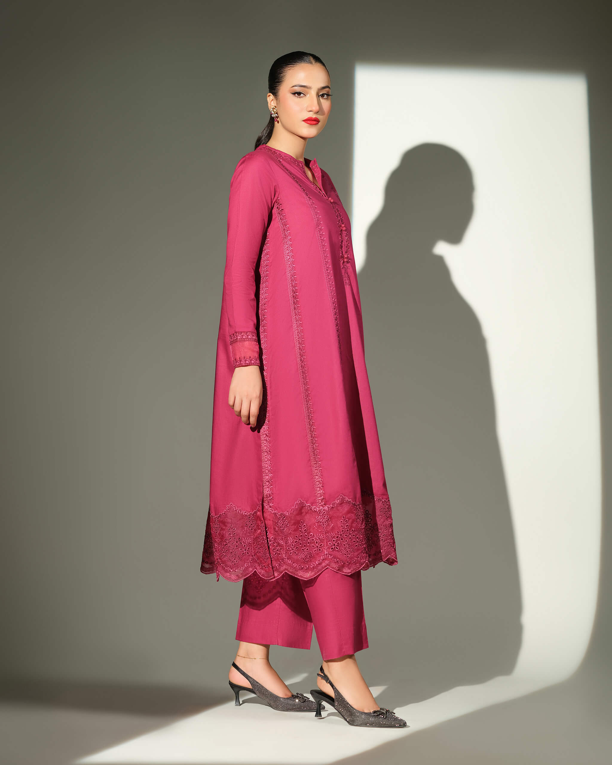 Sinoor Shop pret casual 2025 Sible Dark Pink outfit. A woman walks in profile, wearing a dark pink kurta with detailed embroidery on the hem and sleeves, and matching trousers. She has dark hair pulled back, red lipstick, and is wearing embellished black heels. Full-length shot, studio lighting with a strong shadow.