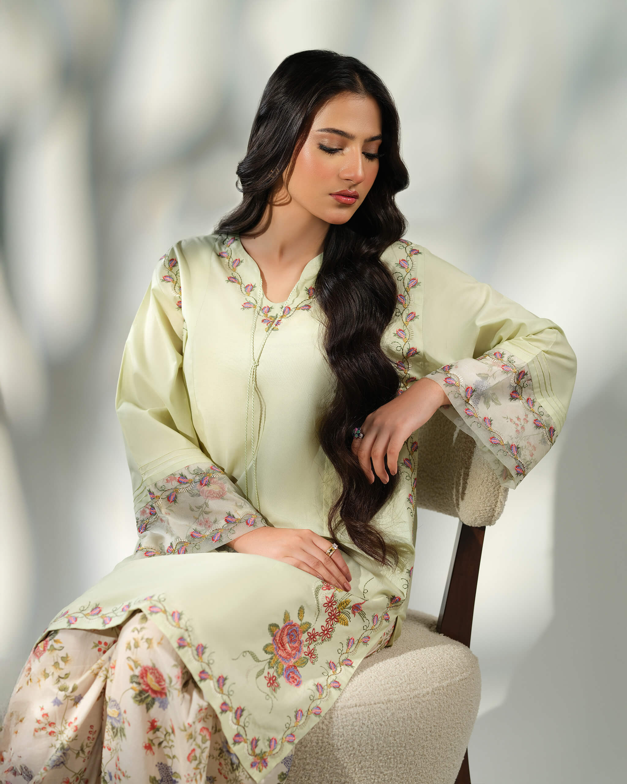 Sinoor Shop pret casual 2025 Zohra Mint Green outfit. A woman with long, dark hair sits in a chair, wearing a light mint green embroidered kurta and floral print trousers. The kurta features embroidery around the neckline and floral print cuffs. Soft, natural lighting.