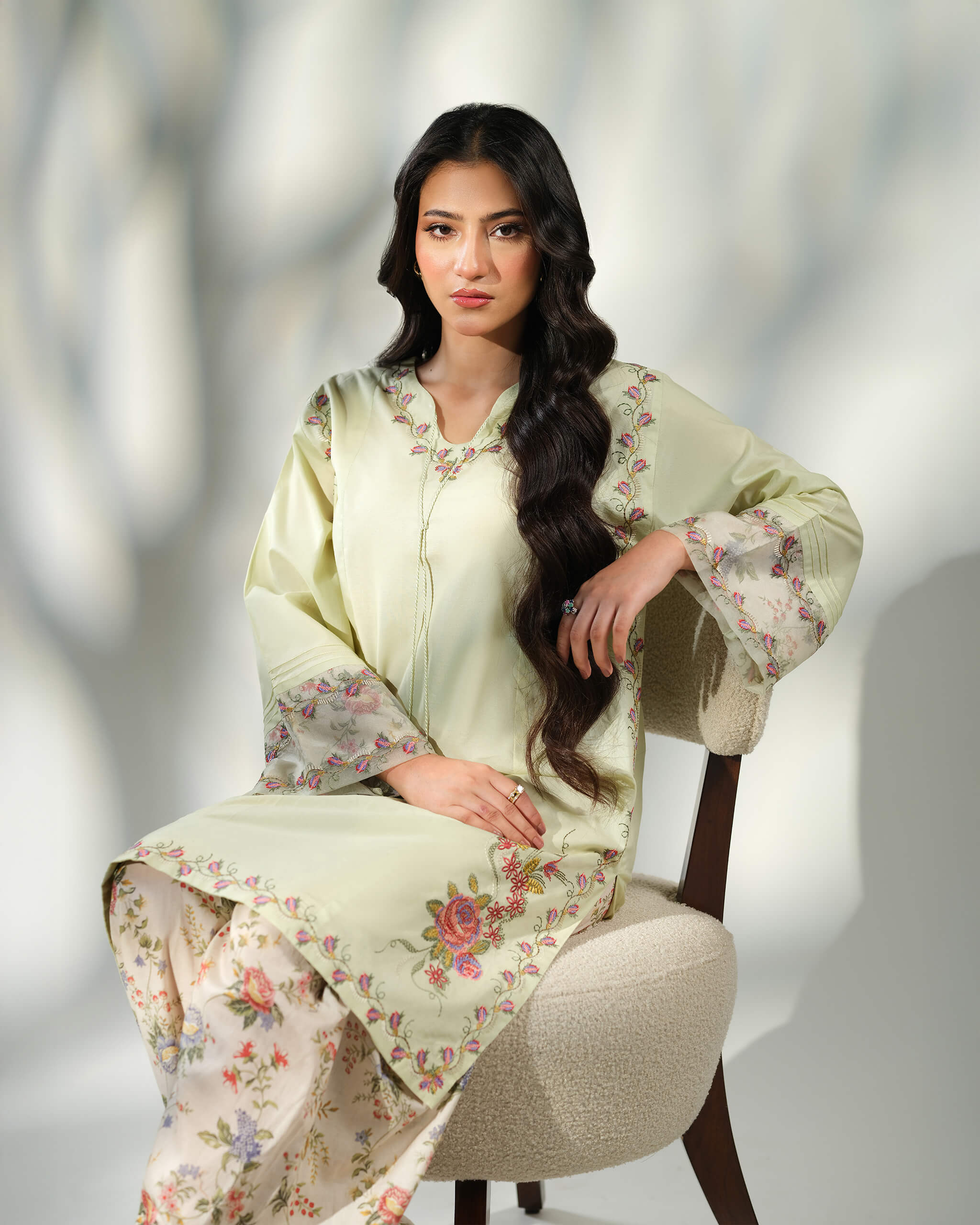 Sinoor Shop pret casual 2025 Zohra Mint Green outfit. A woman with long, dark wavy hair is seated in a chair, modeling a light mint green kurta with floral embroidery and cuffs, paired with floral print trousers. Soft studio lighting with shadows.