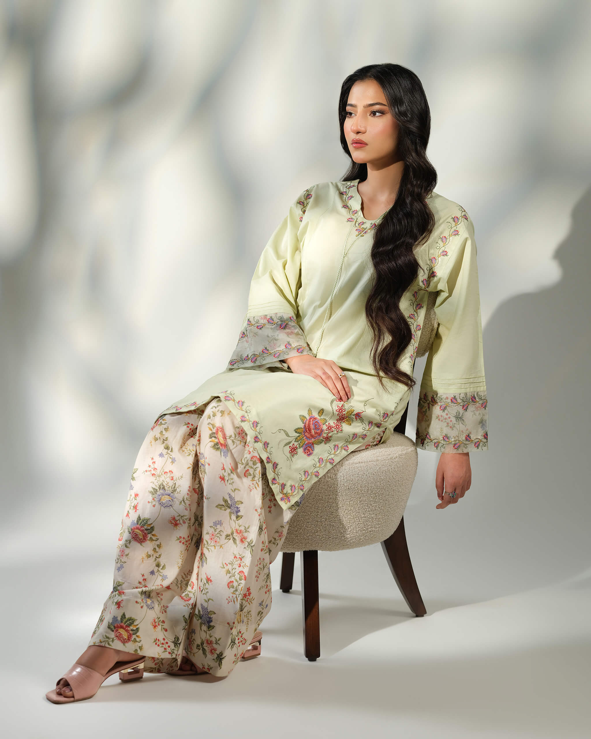 Sinoor Shop pret casual 2025 Zohra Mint Green outfit. A woman with long, dark wavy hair is seated on a stool, wearing a light mint green kurta with floral embroidery and patterned cuffs, along with matching floral print trousers. She is wearing light-colored heels. Soft studio lighting with a shadow.