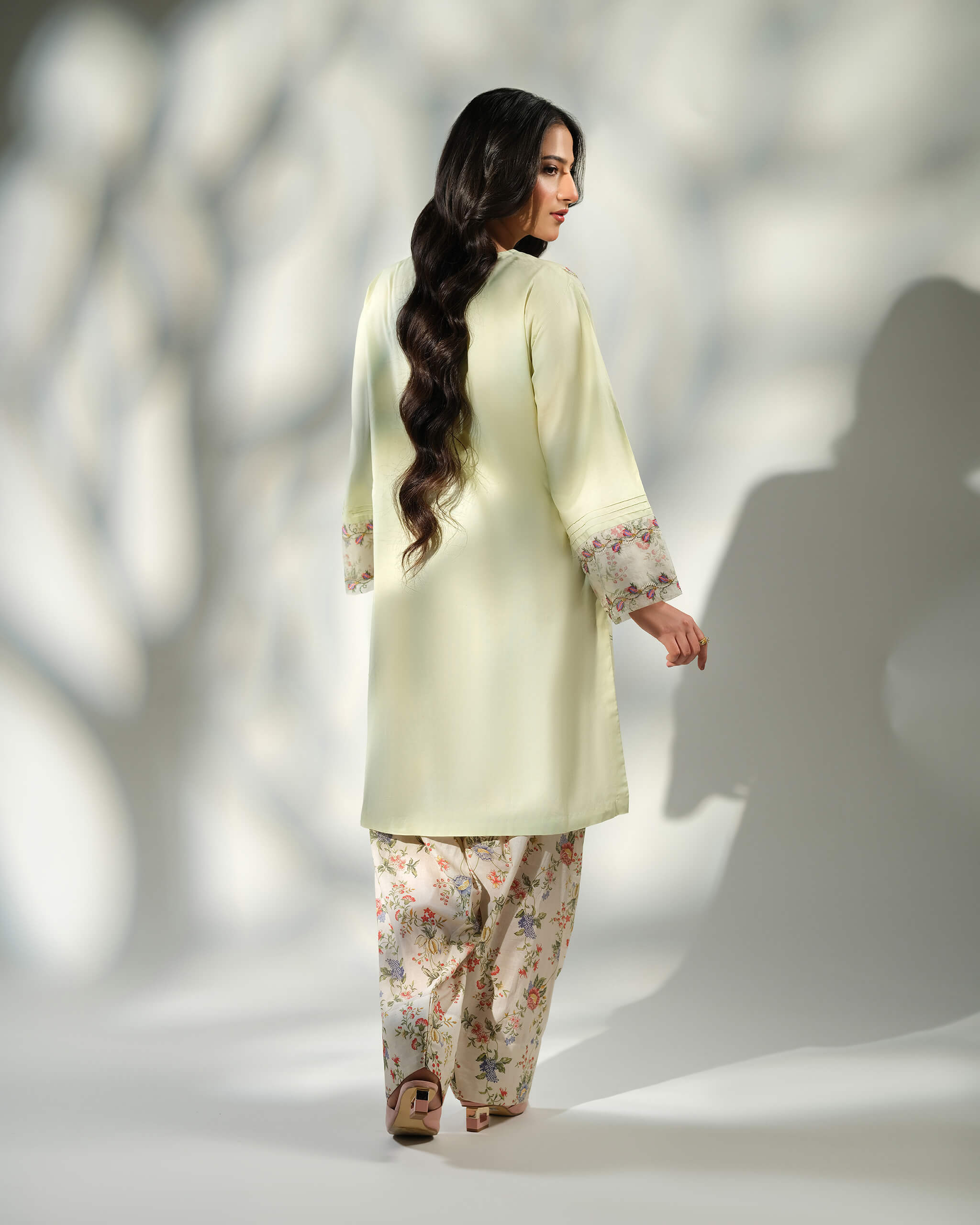 Sinoor Shop pret casual 2025 Zohra Mint Green outfit. A woman with long, dark wavy hair is viewed from the back, modeling a light mint green kurta with floral patterned cuffs and matching floral print trousers. She is wearing light brown heels. Back view, soft studio lighting with shadows.