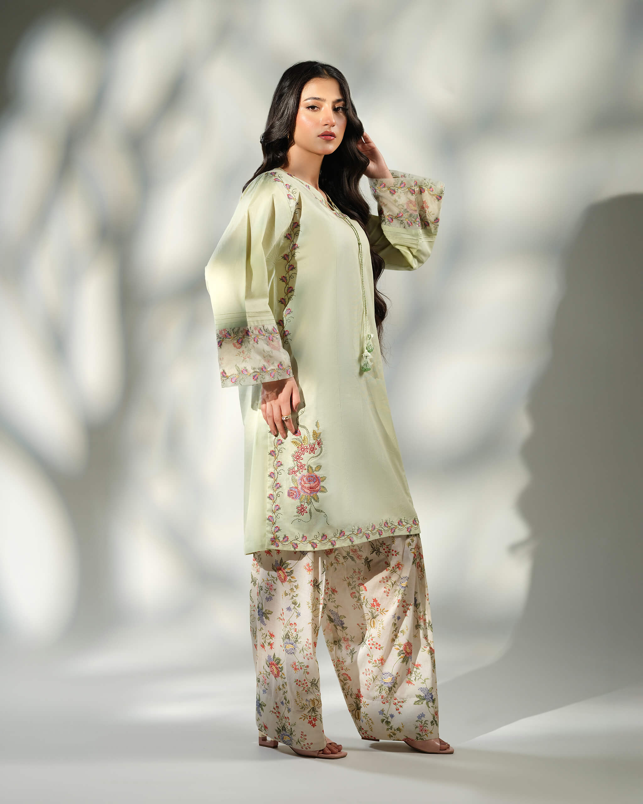 Sinoor Shop pret casual 2025 Zohra Mint Green outfit. A woman stands in a light mint green embroidered kurta with floral print cuffs and matching wide-leg floral print trousers. She has long, dark wavy hair and is wearing light-colored heels. Full-length shot, soft studio lighting with shadows.