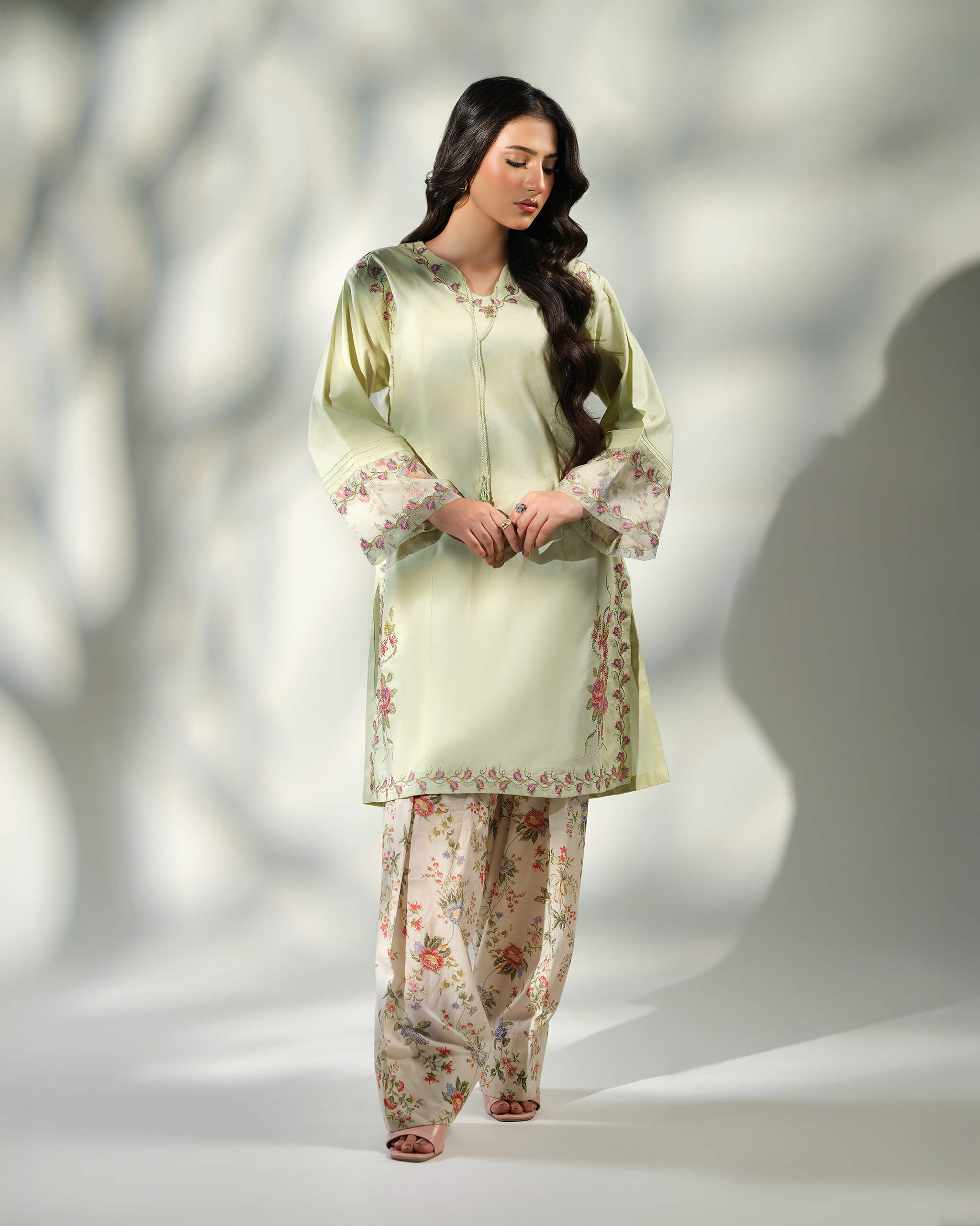 Sinoor Shop pret casual 2025 Zohra Mint Green outfit. A woman stands looking down, wearing a light mint green kurta with floral embroidery and patterned cuffs, paired with matching floral print trousers. She has long, dark wavy hair and is wearing light-colored flat sandals. Full standing shot, soft studio lighting with shadows.